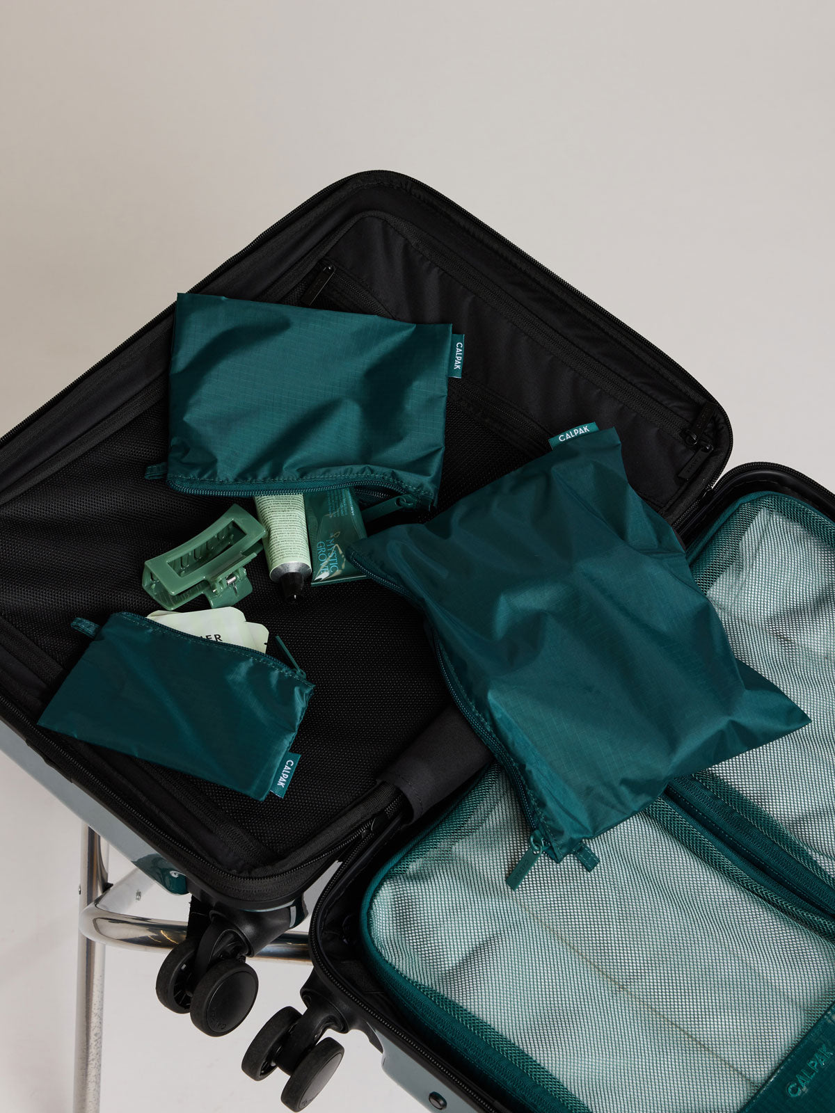 Three dark green teal Kale CALPAK Compakt Zippered pouches of different sizes, arranged inside an open black suitcase, with toiletries visible beneath them on a light neutral background; KZB2001-KALE