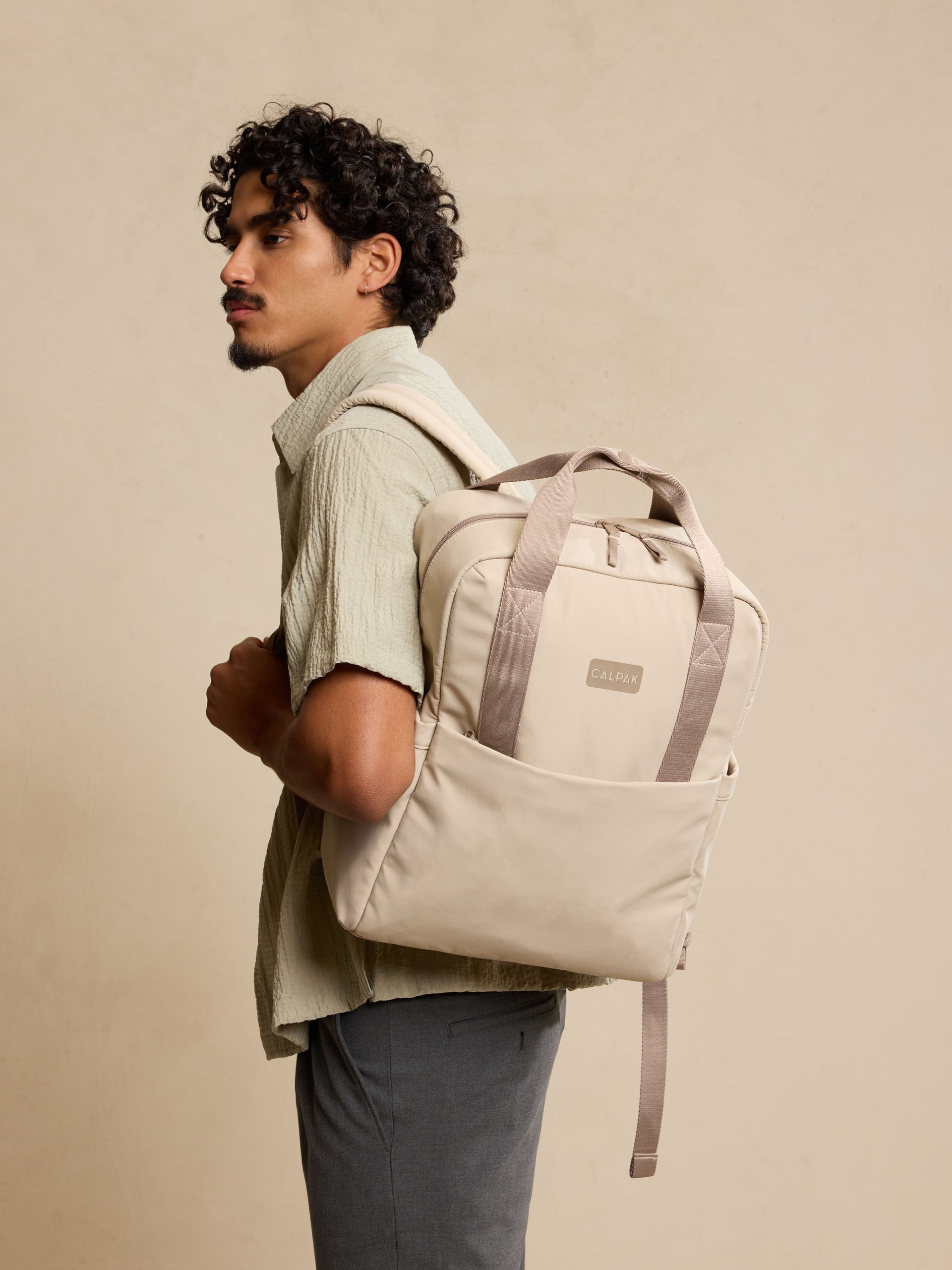 CALPAK Connect Laptop Backpack in grey beige Greige being worn by model  on a beige background; BCN2401-GREIGE