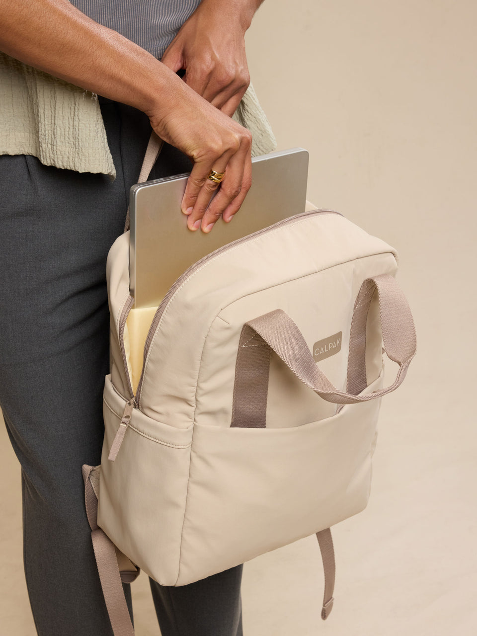 CALPAK Connect Laptop Backpack in grey beige Greige with model putting in laptop into backpack; BCN2401-GREIGE