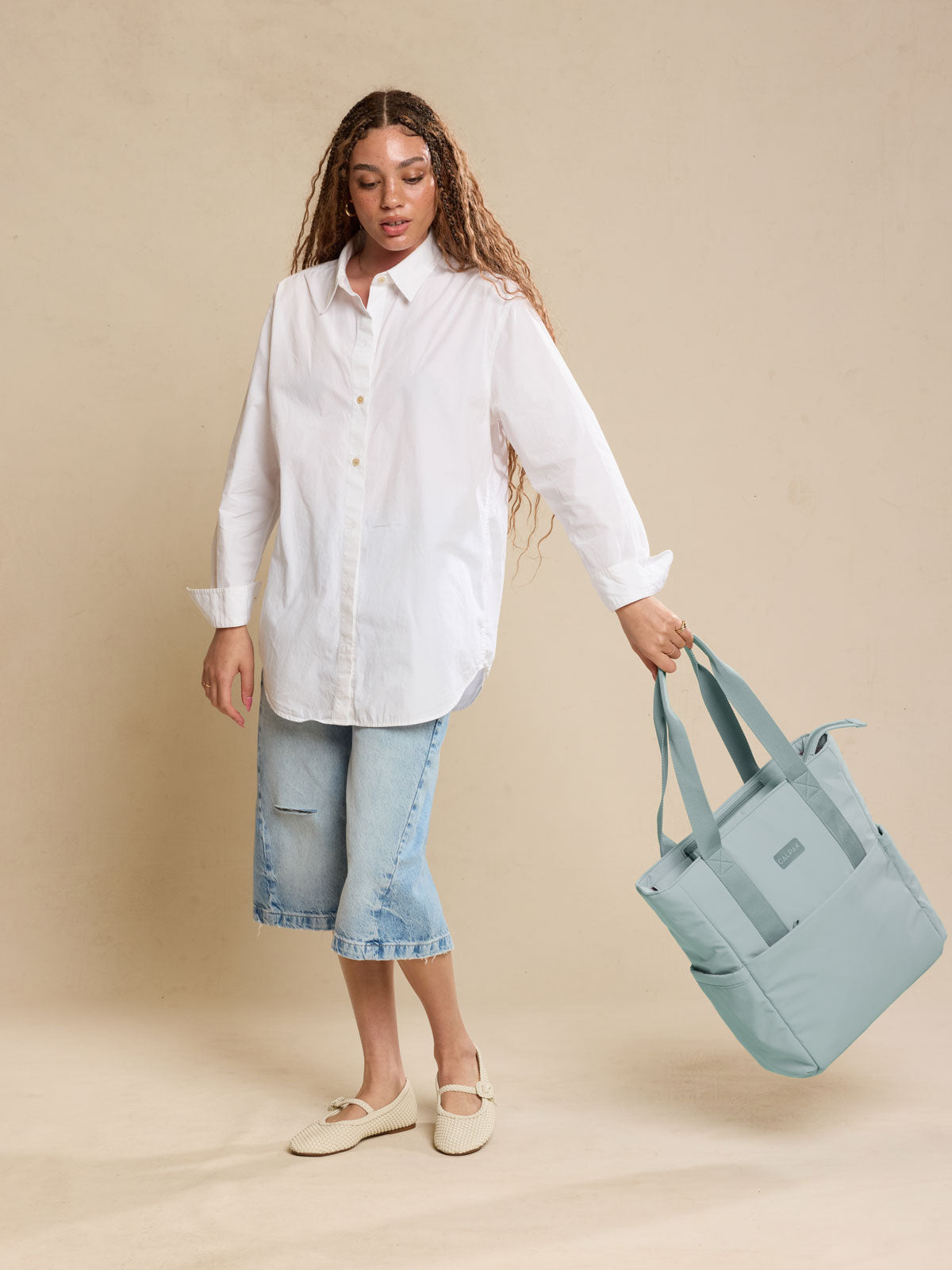 CALPAK Zippered Laptop Tote Bag in light blue Bluebell on a beige background being held by a model; TCN2501-BLUEBELL
