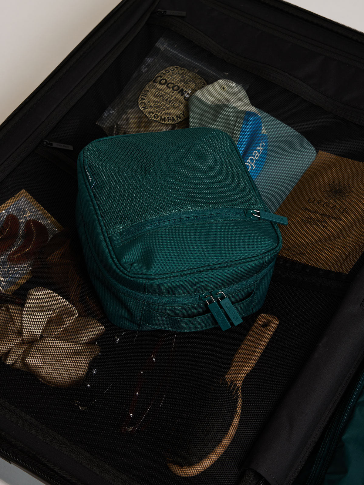 A dark teal green Kale CALPAK Everything Cube organizer  packed inside an open black suitcase, surrounded by travel items including a brown hairbrush, a beige ribbon, and packaged accessories; ACE2401-KALE