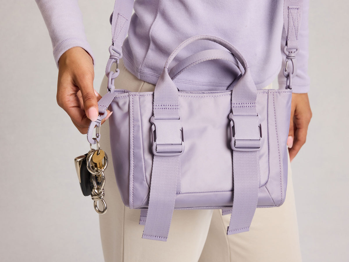 Person holding a light purple CALPAK Fit Gym Crossbody Bag with a matching strap against a neutral background