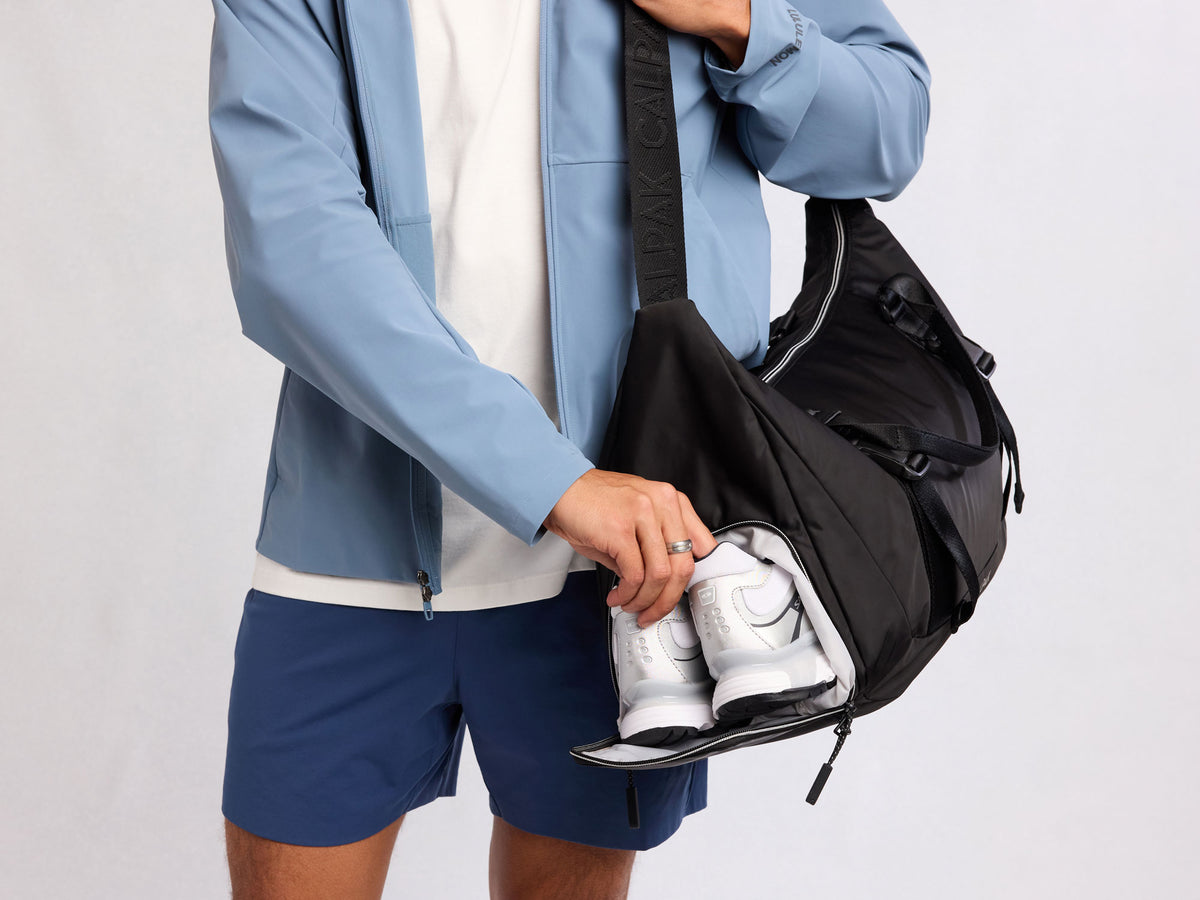 Person holding a black CALPAK Fit Gym duffel bag with white sneakers on a light gray background