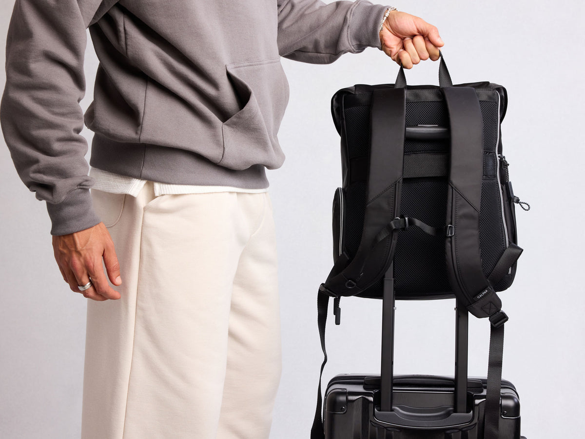 Person holding a black CALPAK Fit Fym Backpack with a suitcase attached on a light gray background