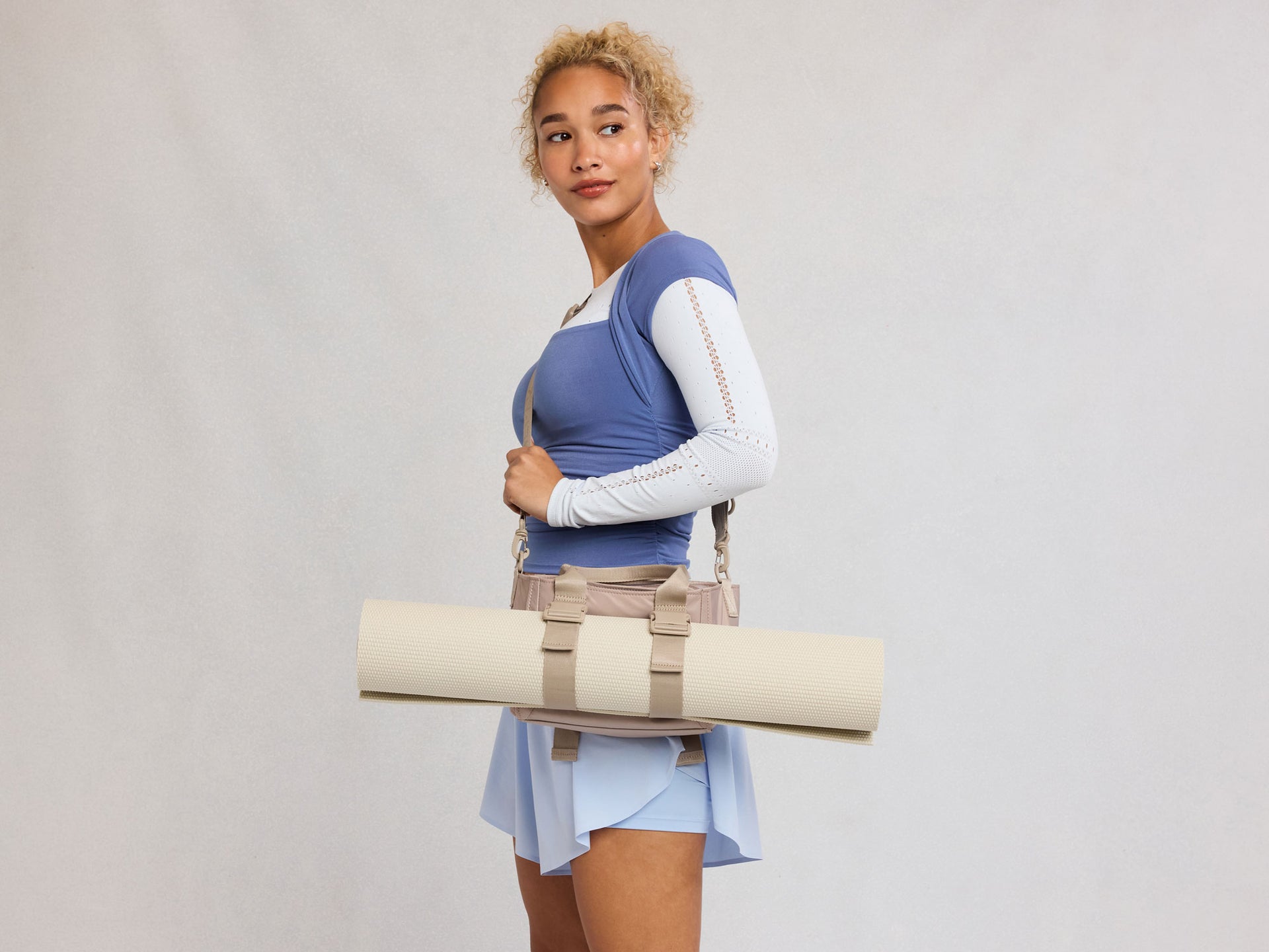 Woman holding a CALPAK Fit Crossbody Bag with yoga mat attached with a neutral background
