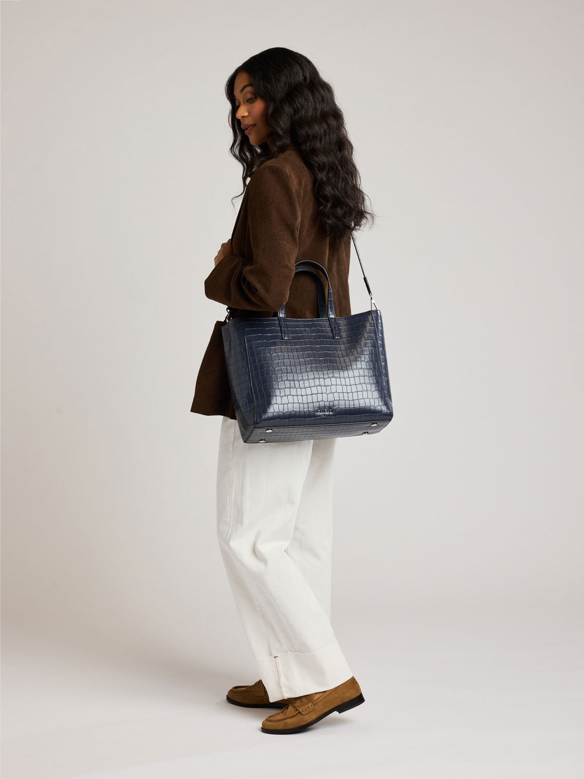 A woman in a brown blazer, white trousers, and brown loafers is shown from the side, carrying a dark blue Navy Haven 15" Laptop Tote in crocodile-embossed, against a plain light gray studio background; ; ATS2501-NAVY-FAUX-CROC