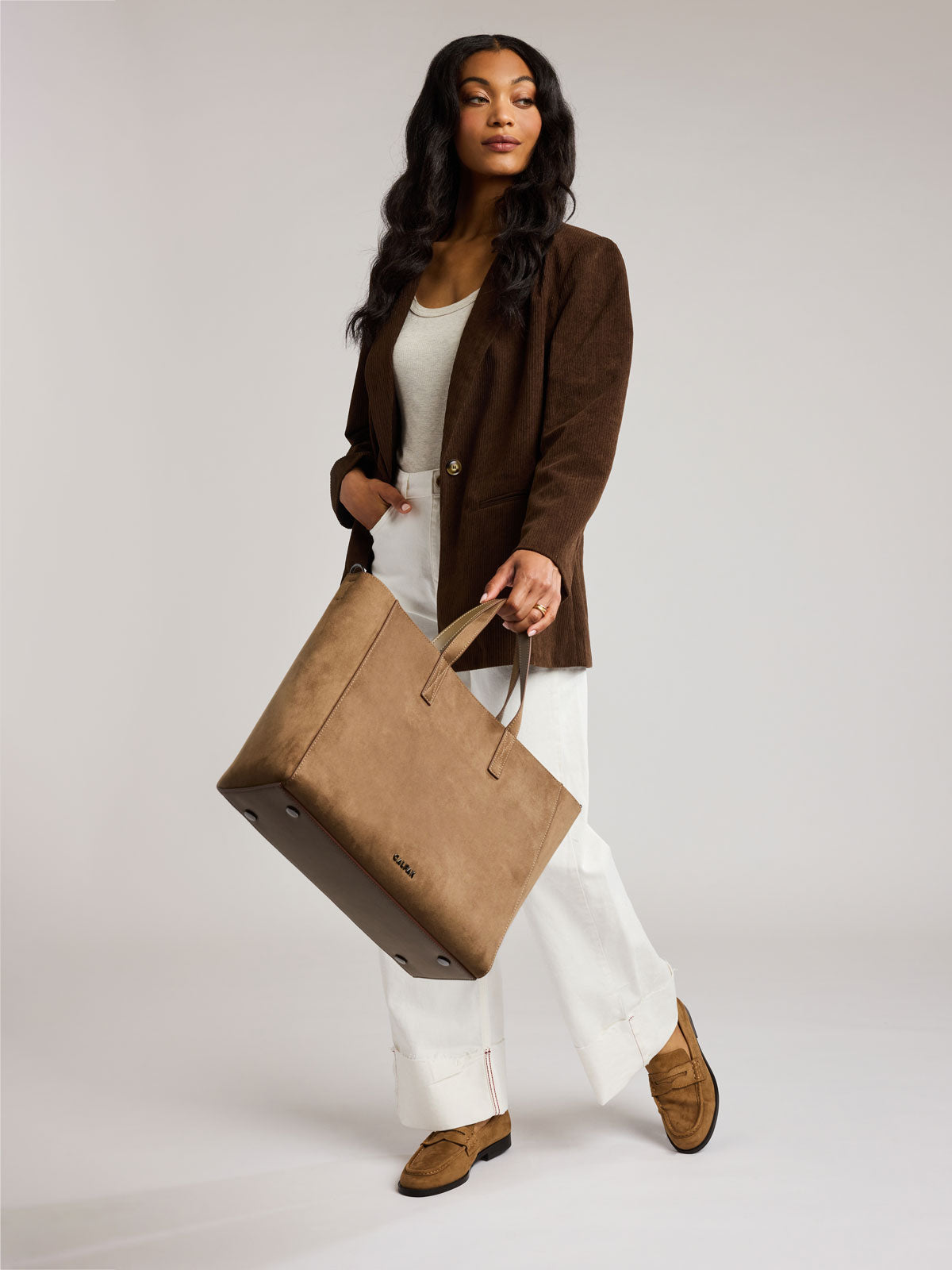 A woman modeling a Haven 15" Laptop Tote in Truffle Suede, styled with a brown blazer, white top and trousers, and brown suede loafers, against a light gray studio background; ATS2501-TRUFFLE-FAUX-SUEDE
