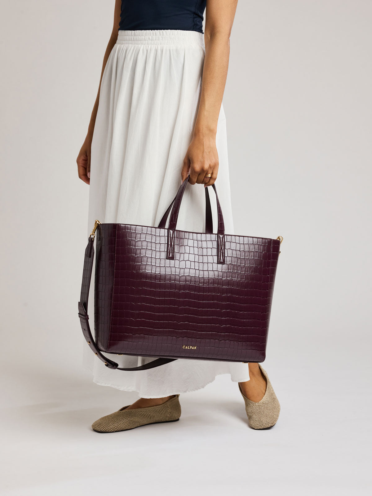 A model wearing a white maxi skirt and beige slip-on shoes holds a large, dark burgundy crocodile-embossed tote bag with gold-tone hardware and a small gold logo on the front; ATO2102-DARK-CHERRY-FAUX-CROC