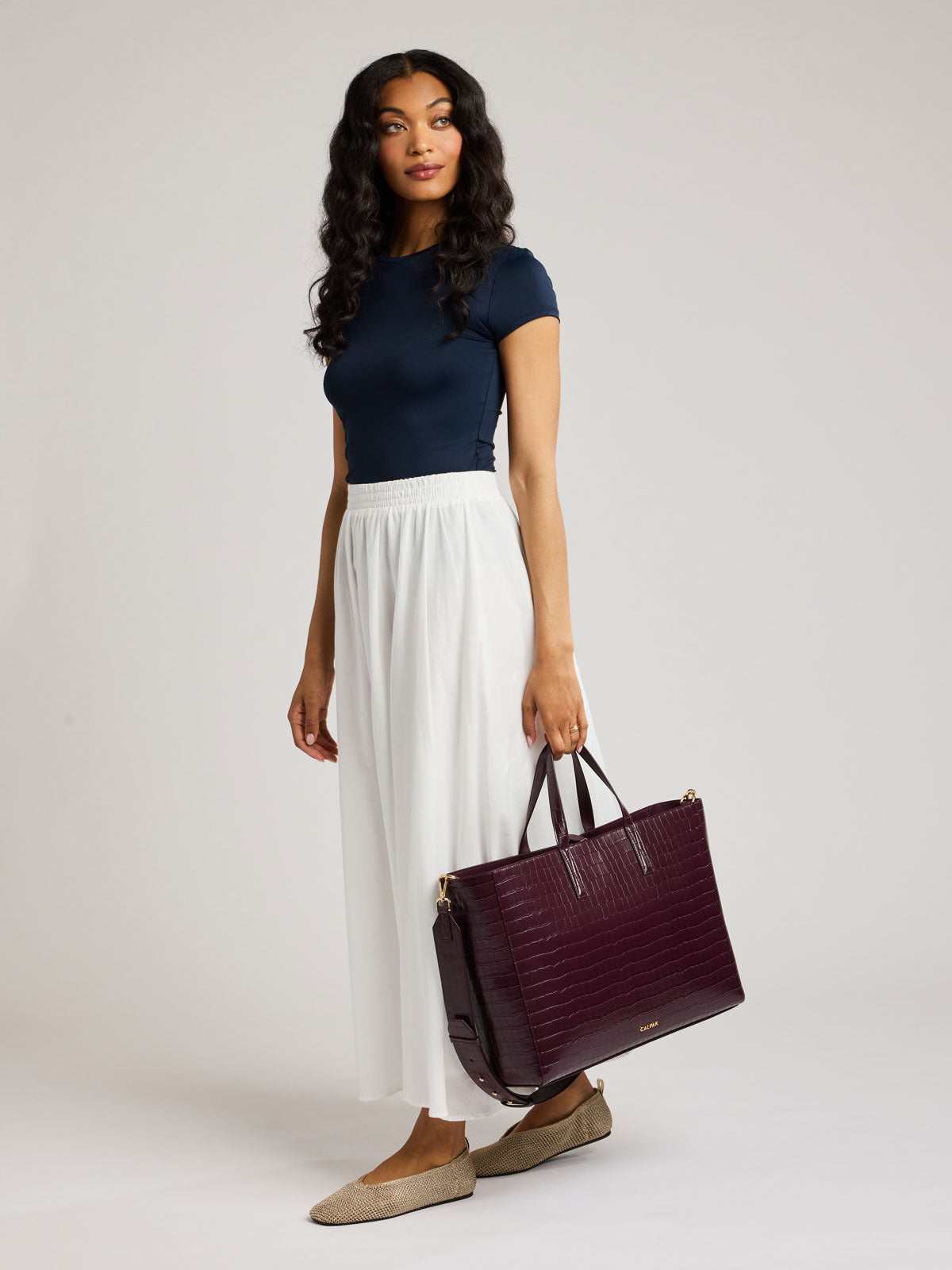 A woman modeling a navy blue short-sleeve top, white pleated maxi skirt, and beige flats, holding a dark burgundy crocodile-embossed tote bag against a light gray studio background; ATO2102-DARK-CHERRY-FAUX-CROC
