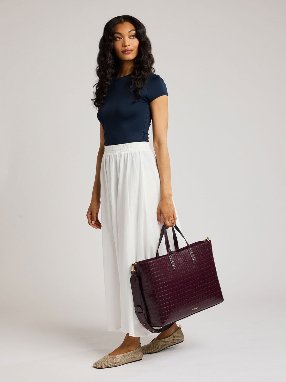 A woman modeling a navy blue short-sleeve top, white pleated maxi skirt, and beige flats, holding a dark burgundy crocodile-embossed tote bag against a light gray studio background; ATO2102-DARK-CHERRY-FAUX-CROC
