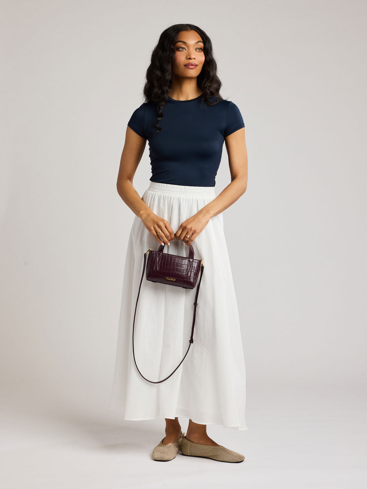 A woman modeling a navy blue short-sleeve top and a white midi skirt, holding a small Haven Mini Crossbody Bag in Dark Cherry against a light gray studio background; AMC2502-DARK-CHERRY-FAUX-CROC