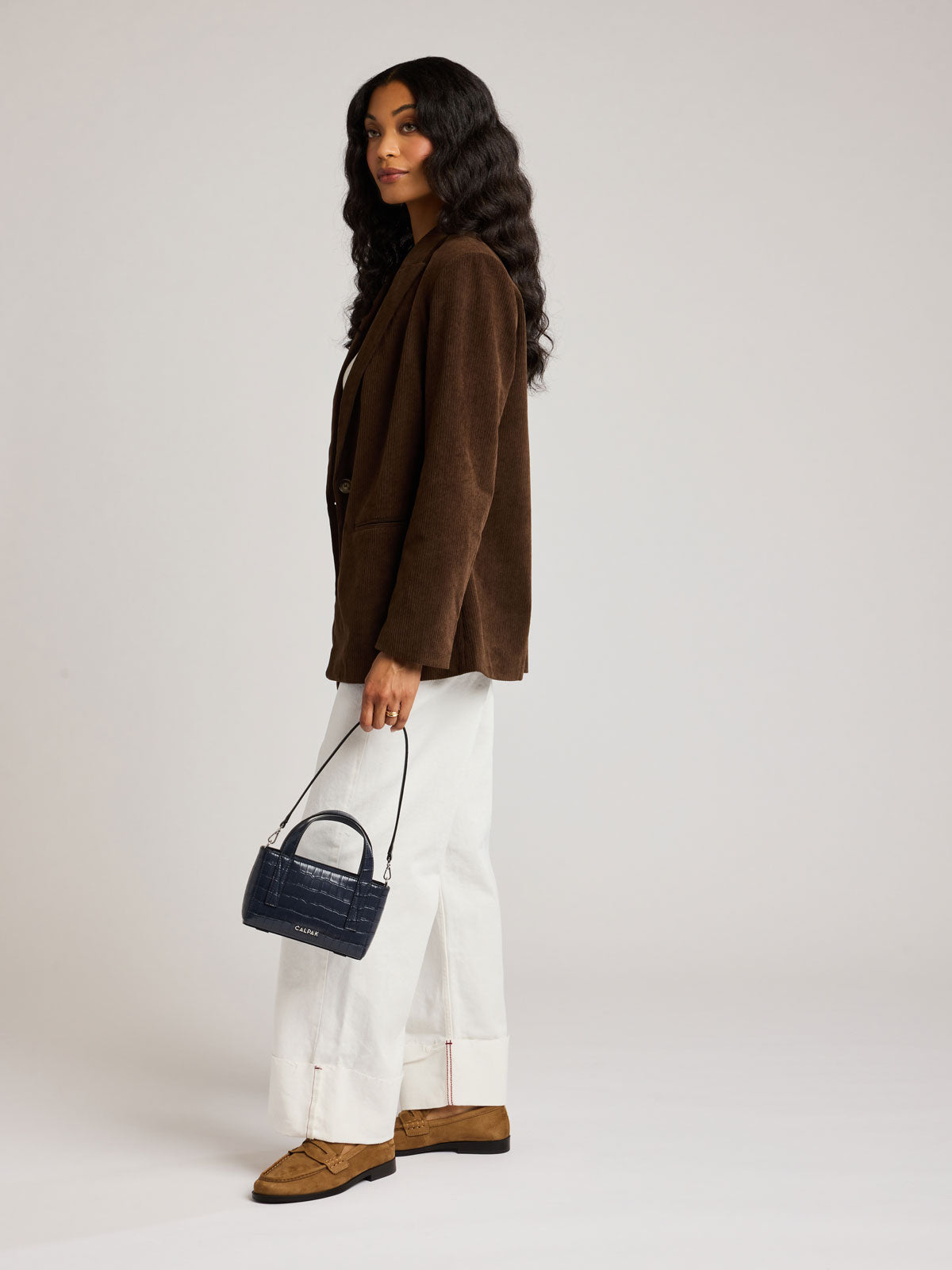 A woman stands in profile wearing a brown corduroy blazer, white wide-leg trousers, and brown suede loafers, while holding a Haven Mini Crossbody Bag in Navy dark blue croc-embossed handbag against a plain light gray studio background; AMC2502-NAVY-FAUX-CROC