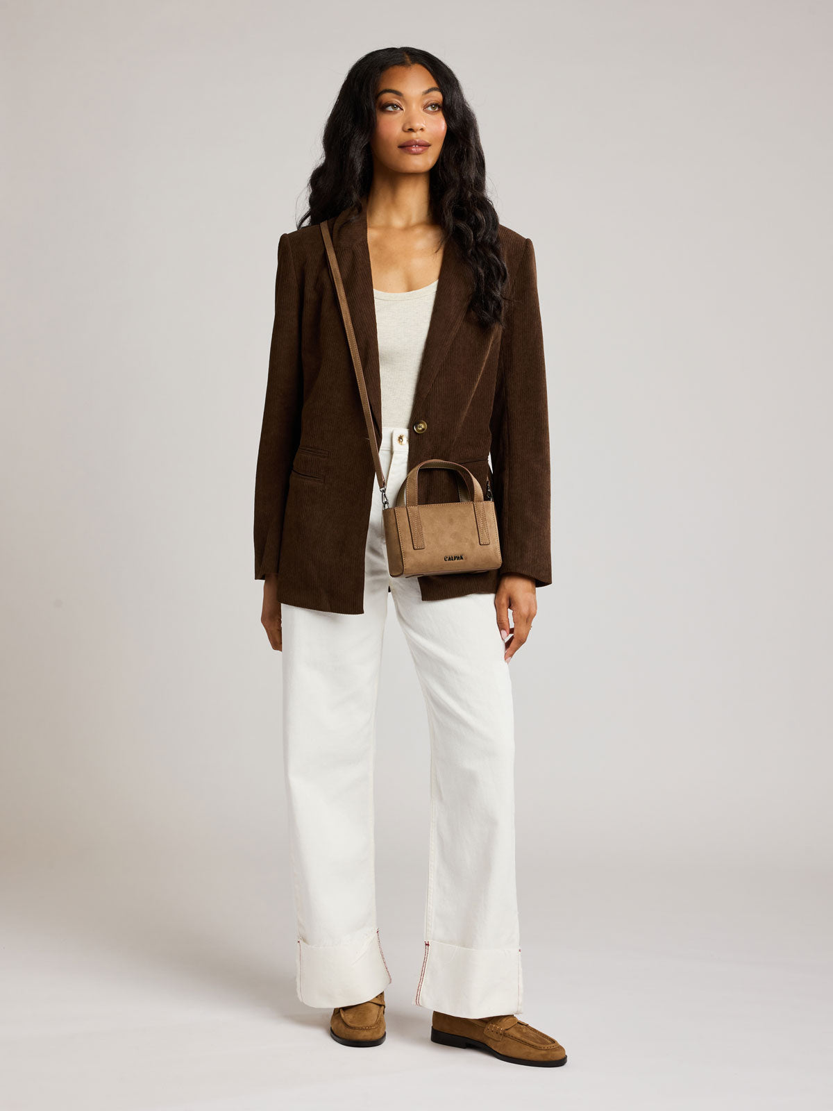 A woman stands against a light gray studio background wearing a brown corduroy blazer, white top and wide-leg white pants, with a small Haven Mini Crossbody Bag in tan brown Truffle Suede; AMC2502-TRUFFLE-FAUX-SUEDE