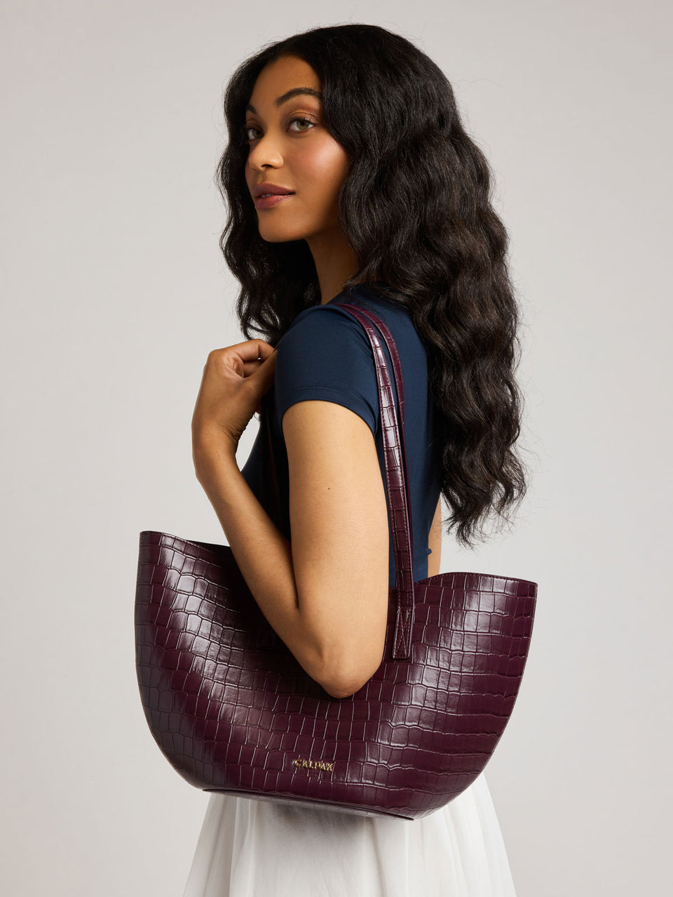 A woman with long, wavy dark hair is shown from the side/back, wearing a Haven Tulip Tote in Dark Cherry that is crocodile-embossed over her shoulder against a neutral light gray studio background; AHP2601-DARK-CHERRY-FAUX-CROC