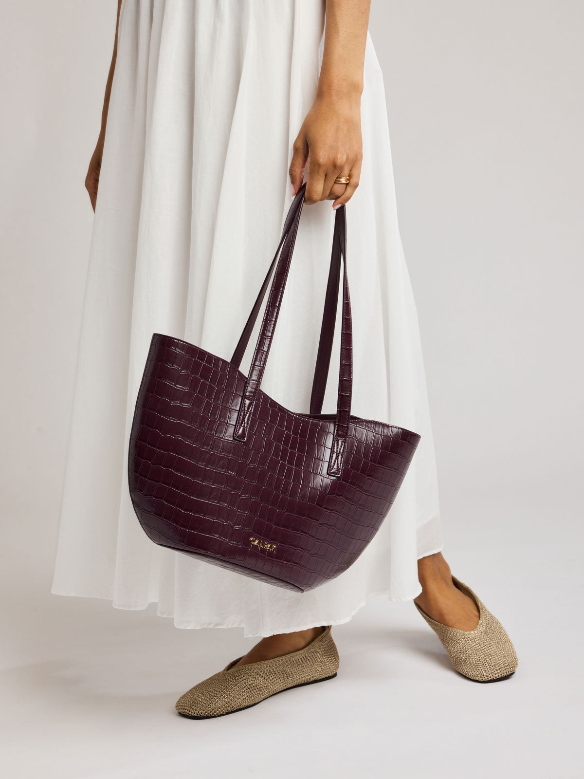 A person wearing a white maxi dress and beige flat shoes holds a large, Haven Tulip Tote in Dark Cherry in crocodile-embossed tote bag with double shoulder straps; AHP2601-DARK-CHERRY-FAUX-CROC