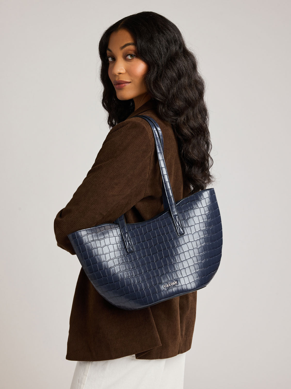 A woman with long, wavy dark hair is shown from the side/back wearing a brown suede jacket and white pants, carrying a Haven Tulip Tote in Navy blue crocodile-embossed tote bag over her shoulder; AHP2601-NAVY-FAUX-CROC