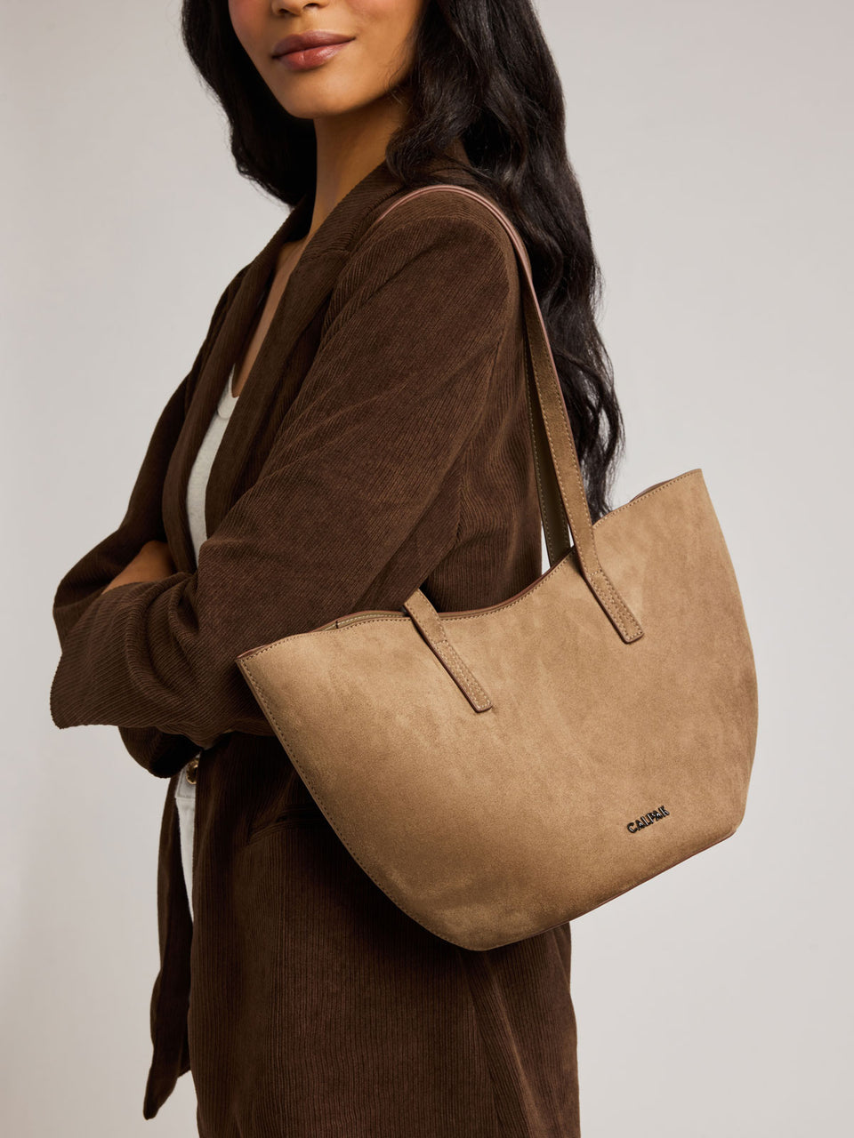 A woman wearing a brown corduroy cardigan carries a Haven Tulip Tote bag in brown tan Truffle Suede with a set against a plain light gray studio background;  AHP2601-TRUFFLE-FAUX-SUEDE