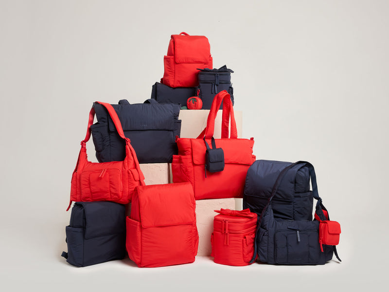 Collection of red and navy CALPAK Baby Collection bags on a light gray background