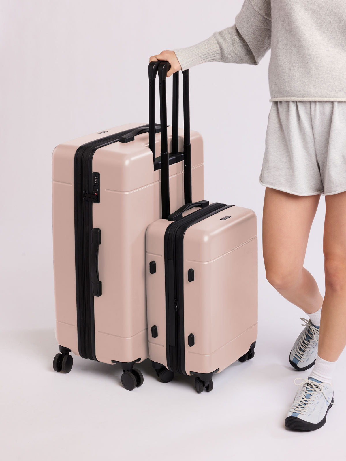 Person holding a set of rose quartz CALPAK luggage part of Hue Starter Bundle with black handles and wheels on a light gray background; LHU8000-ROSE-QUARTZ