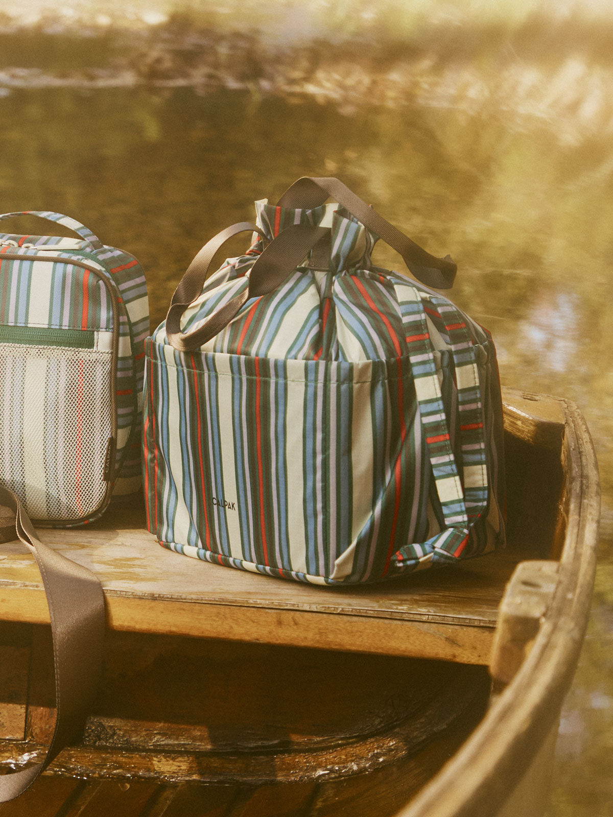 CALPAK Insulated Lunch Bag in multicolor (celadon, mushroom, french blue, lavender) striped Riviera Stripe on a bench; ALB2001-RIVIERA-STRIPE