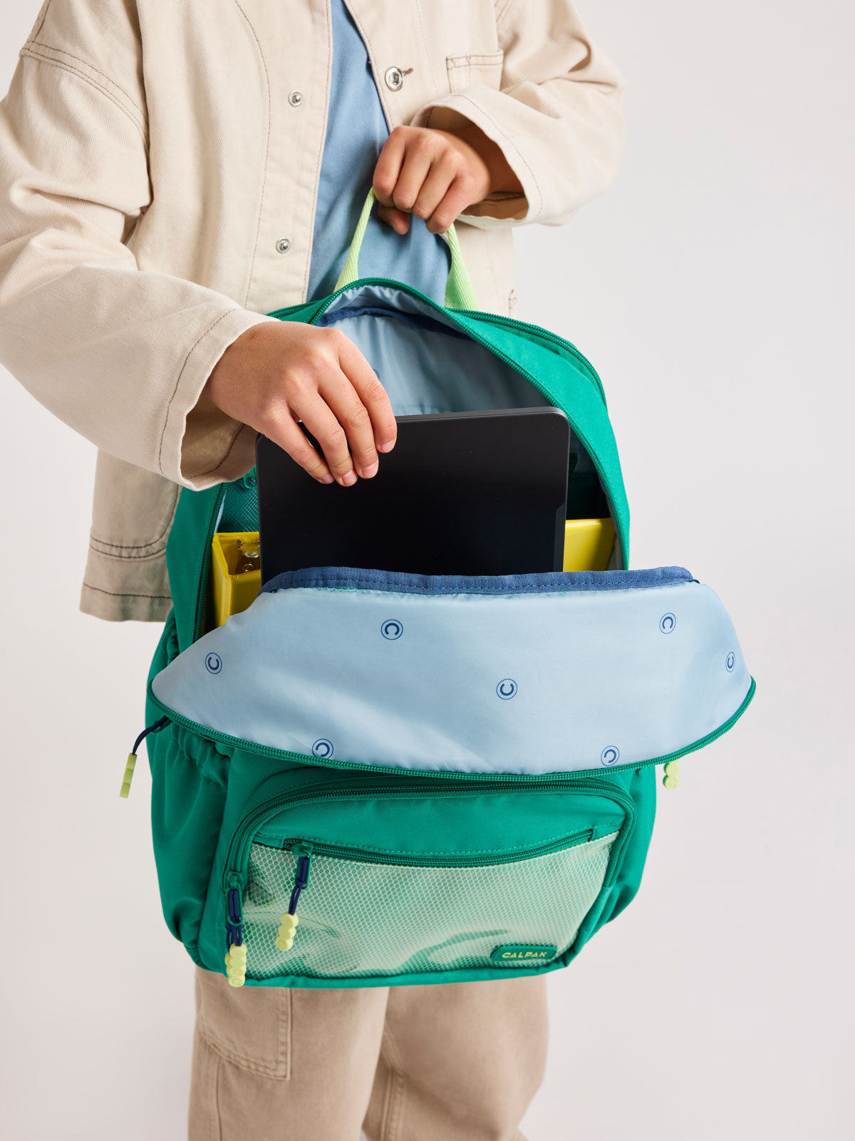 Child putting tablet into Green CALPAK Kids Large Backpack with lime green accents on a white background; CKBL2501-GREEN