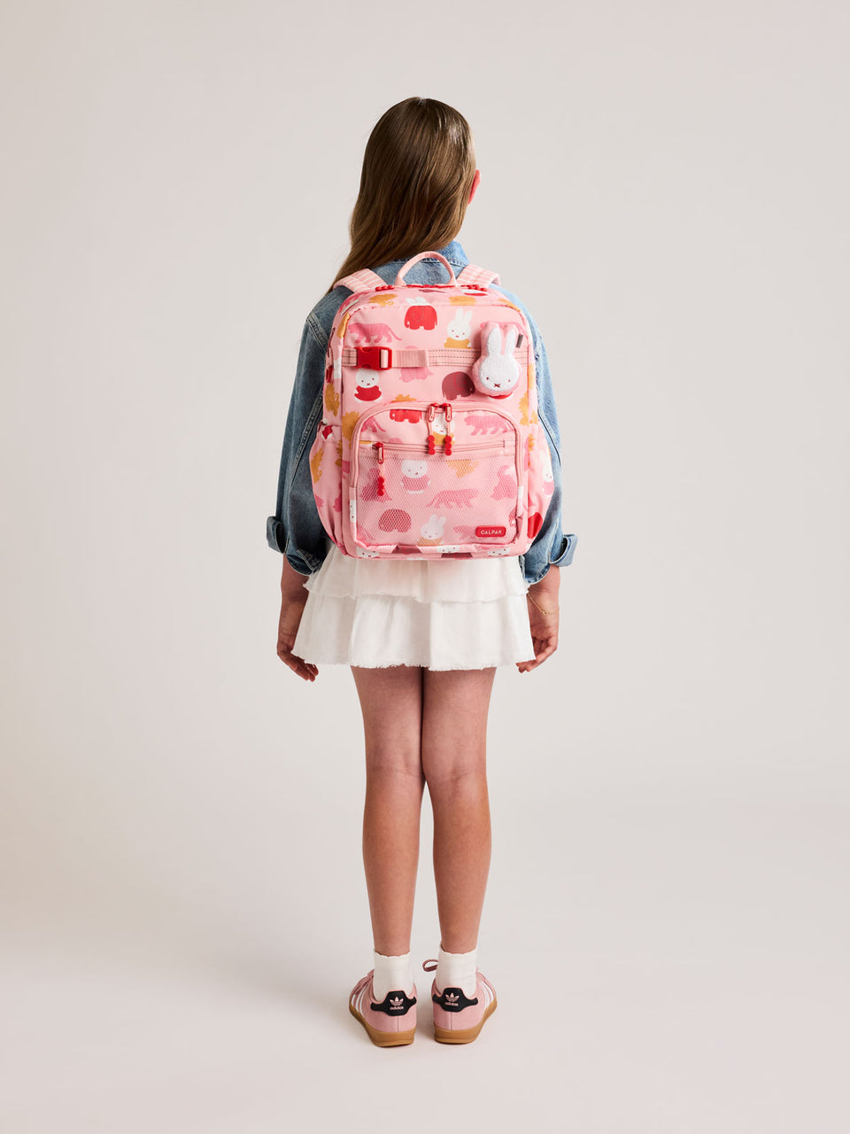 Girl wearing a CALPAK Kids Large Backpack with Miffy and pink colorful animal pattern on a white background; CKBL2501-MIFFY-PINK-ZOO-TRIP