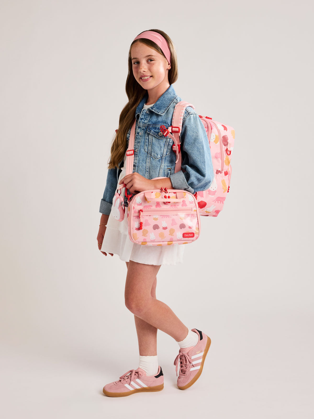 Girl wearing the Pink CALPAK Kids Lunch Bag with Miffy and animal pattern with mesh front pocket and top handles and the Miffy Kids Backpack on a white background; CKLB2501-MIFFY-PINK-ZOO-TRIP