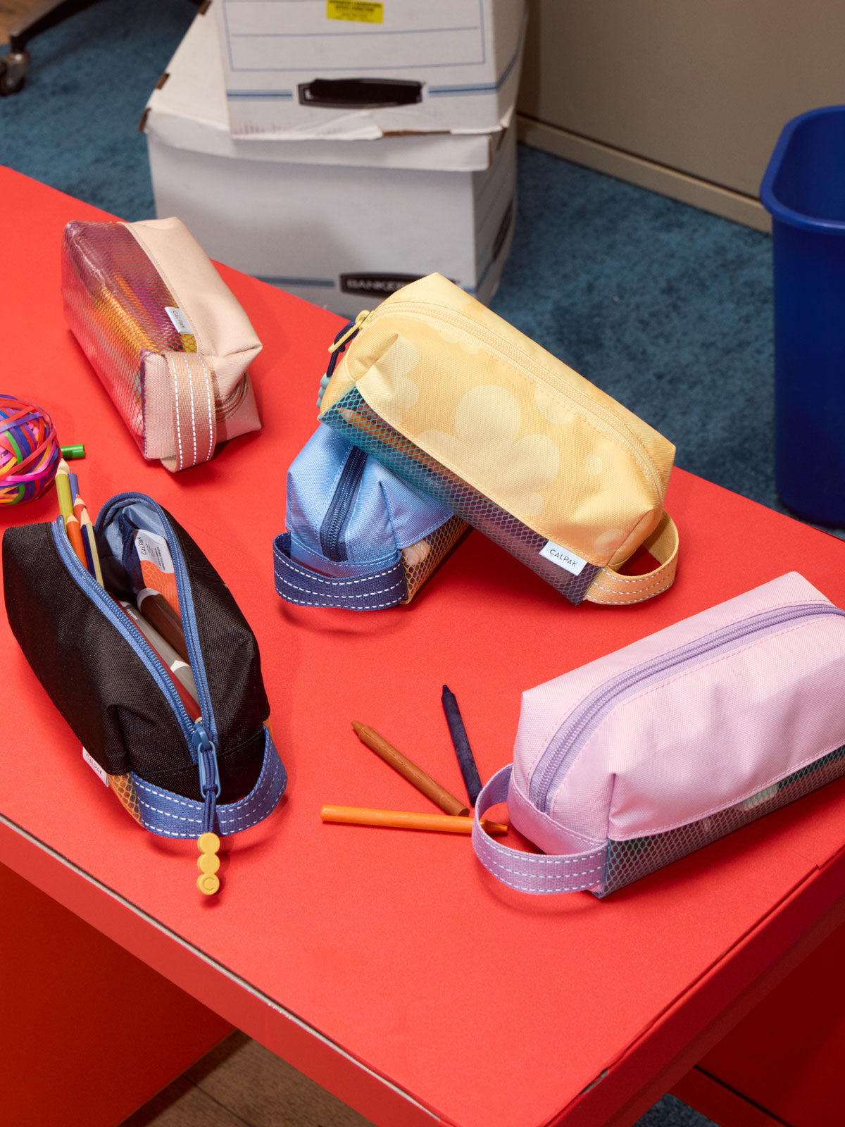 A collection of CALPAK Kids Pencil cases pencil cases arranged on a bright red table, with pens placed in front of them; CKPC2501-BLUE