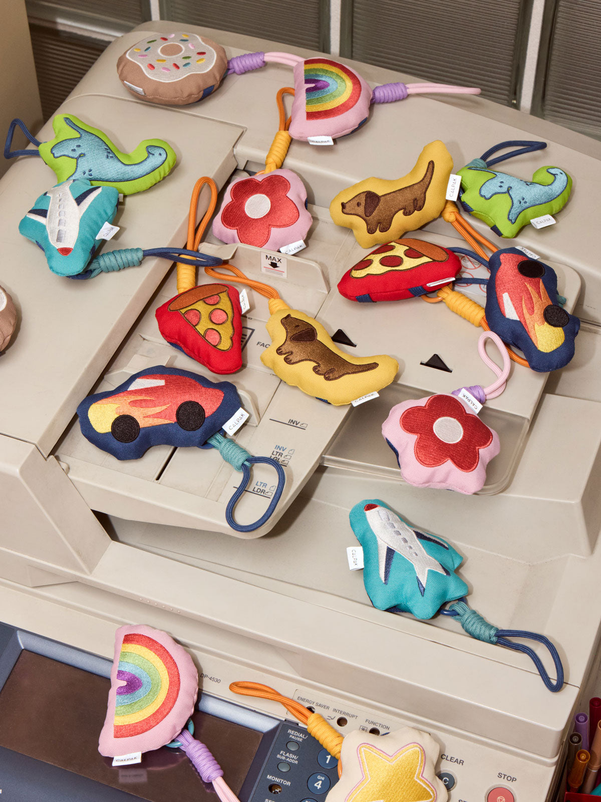 A collection of colorful CALPAK Pluffy Bag Charms shaped like cars, flowers, dinosaurs, a pizza slice, a rocket ship, and rainbows, arranged on top of a beige photocopier; APLF2501-DONUT