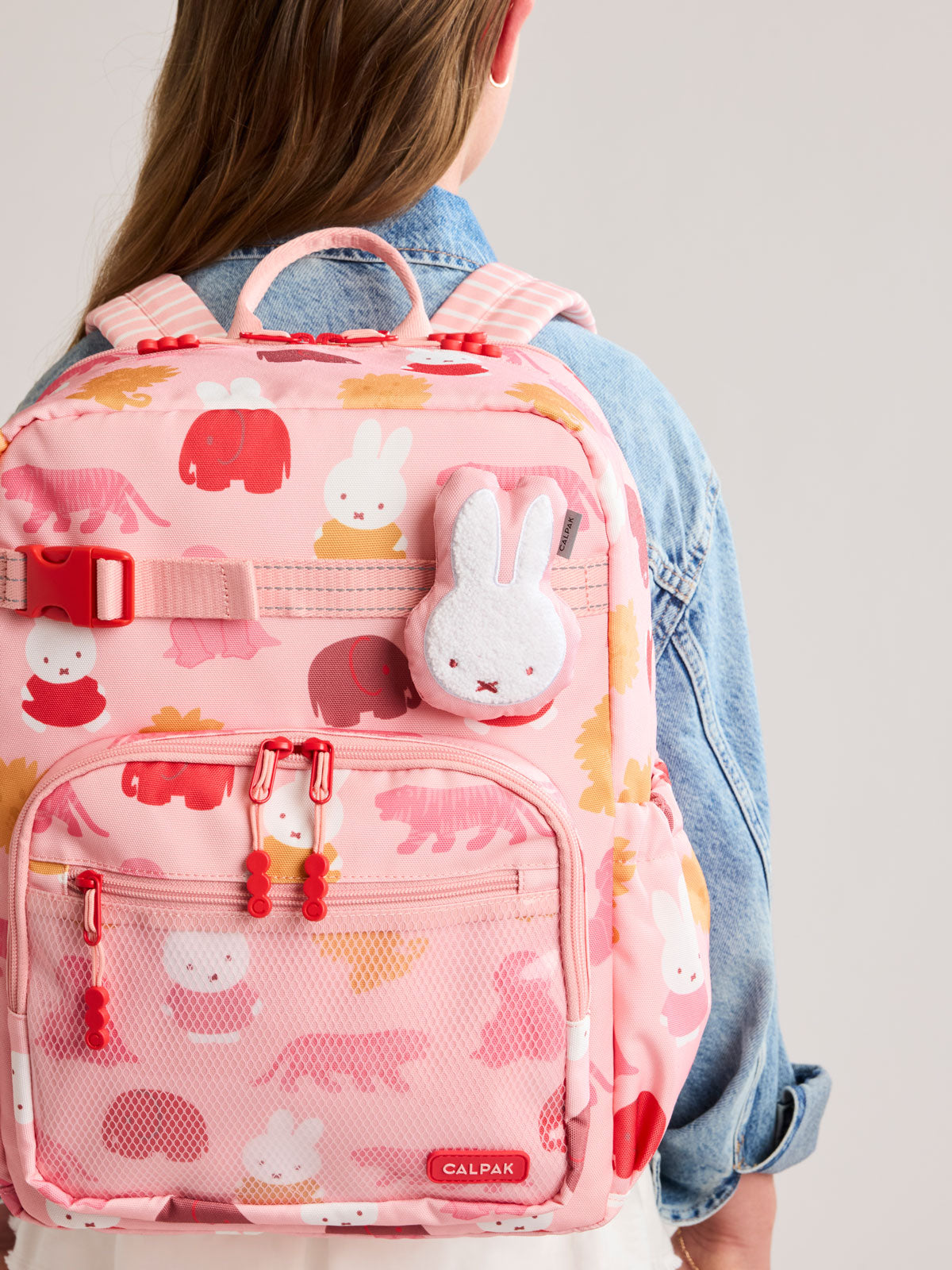 Plush Miffy x CALPAK Pluffy Bag Charm keychain with a pink border and Miffy being worn by a child with a colorful Kids CALPAK Kids Backpack; APLF2501-PINK-MIFFY