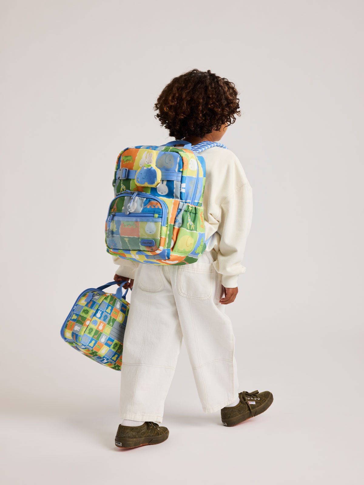 Child wearing the CALPAK Kids Small Backpack with Miffy and blue colorful animal pattern on a white background; CKBS2501-MIFFY-BLUE-ZOO-TRIP