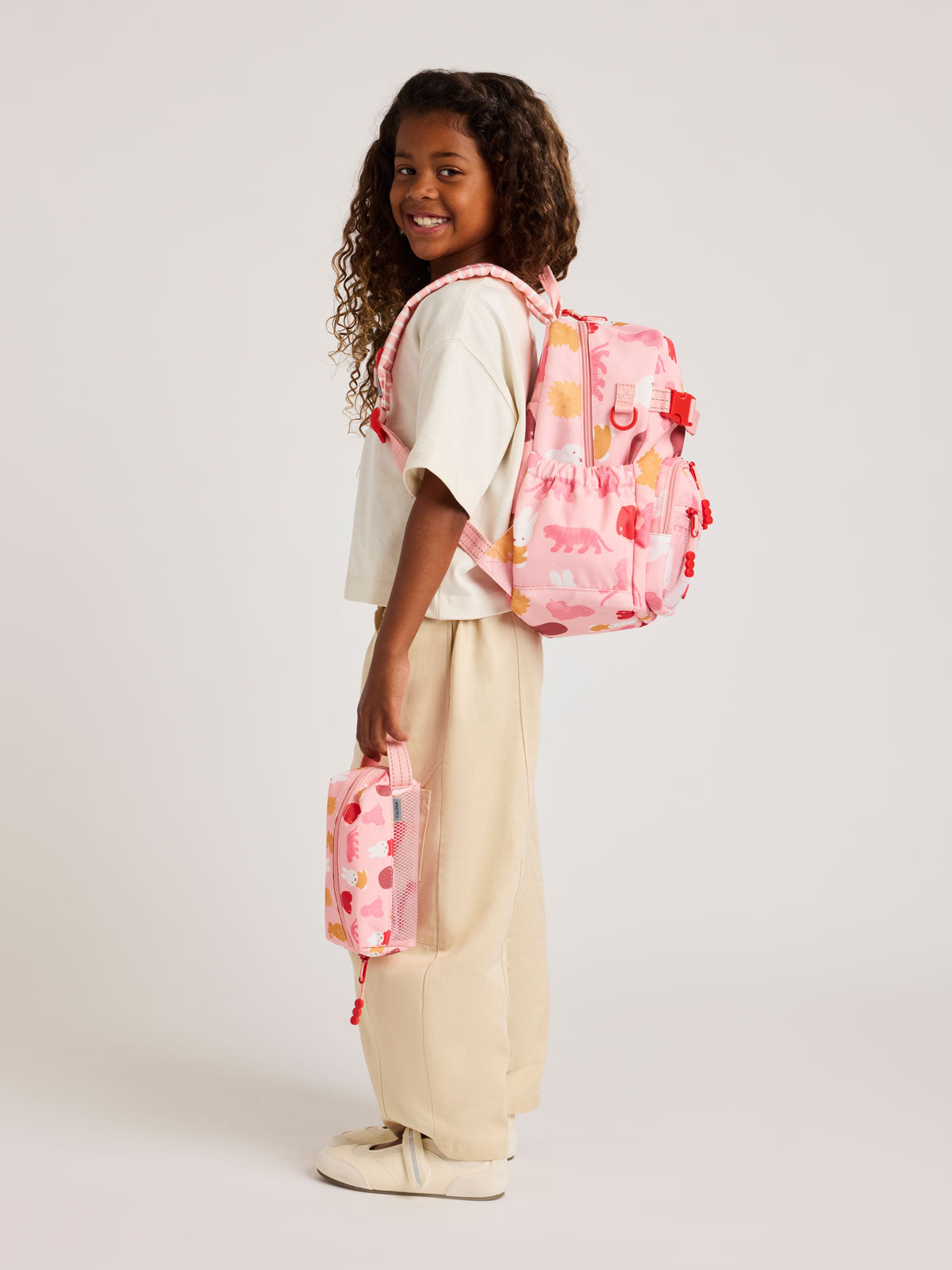 Child wearing a CALPAK Kids Small Backpack with Miffy and pink colorful animal pattern on a white background; CKBS2501-MIFFY-PINK-ZOO-TRIP