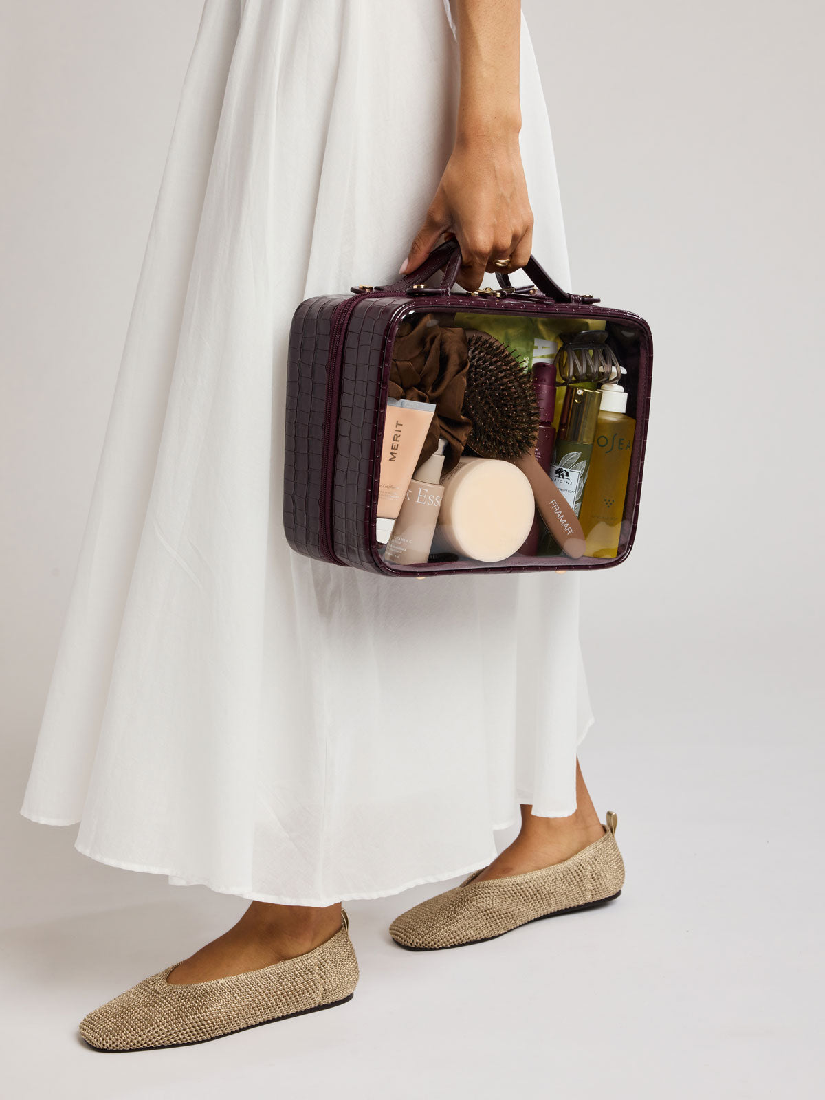 A person in a flowing white dress holds  a CALPAK Large Clear Cosmetics Case in deep red Dark Cherry against a white background; CCC2001-DARK-CHERRY-FAUX-CROC