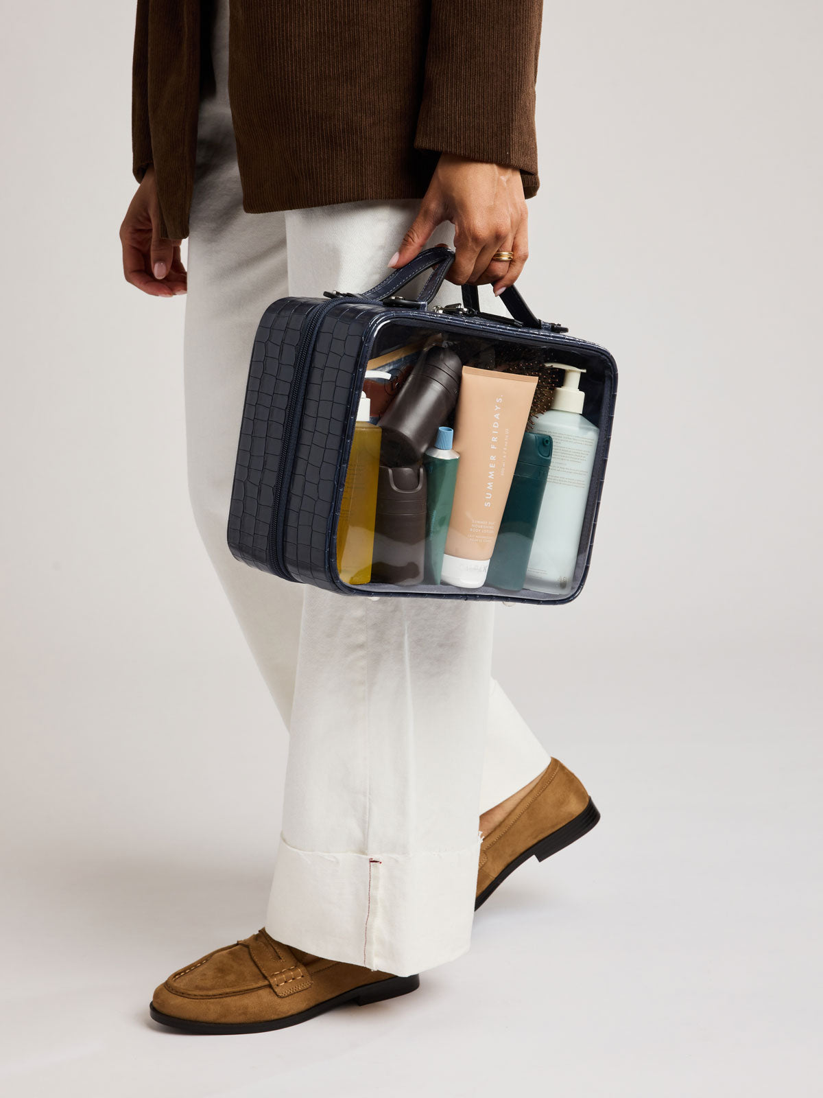 A person wearing a brown jacket, white trousers, and tan loafers carries CALPAK Large Clear Cosmetics Case in deep blue Navy croc-embossed against a white background; CCC2001-NAVY-FAUX-CROC