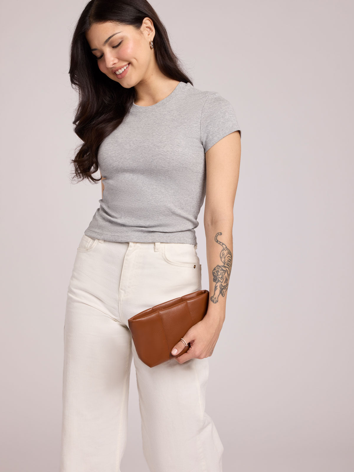 A woman wearing a light gray short-sleeve crewneck t-shirt and off-white wide-leg jeans, holding a CALPAK Luka 1 Quart Pouch in Honey Brown light gray studio background;  ZLPS2601-HONEY-BROWN-PU