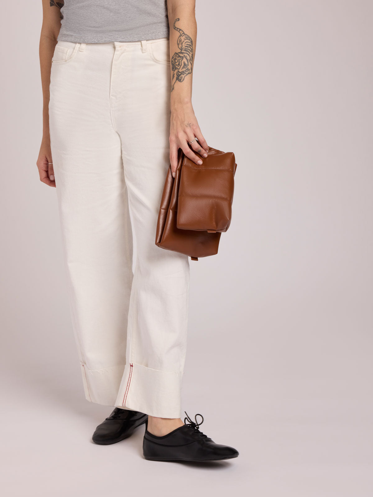 A model wearing off-white cropped trousers with rolled cuffs, black shoes, and holding a CALPAK Luka 2 Quart Pouch in Honey Brown Vegan Leather against a light gray studio background; ZLPM2601-HONEY-BROWN-PU