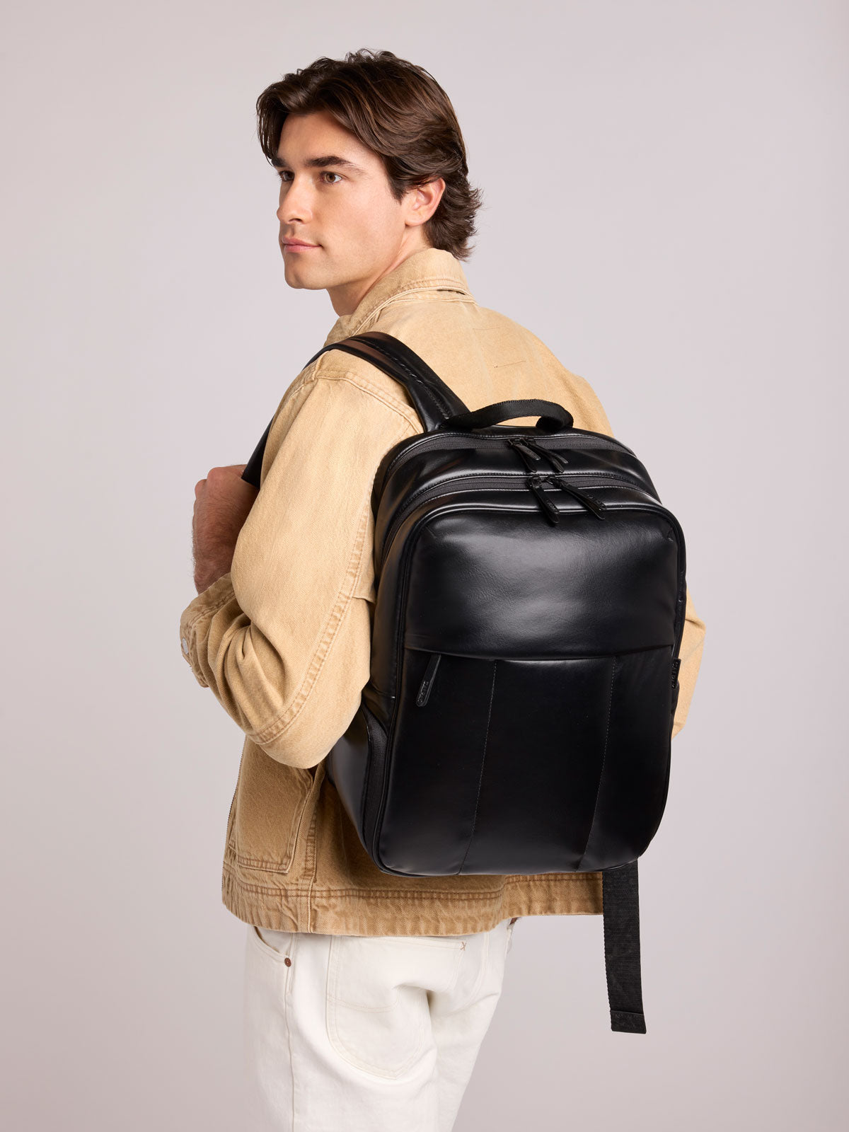 A man wearing a beige jacket and white pants is shown from the back and side, carrying a CALPAK Luka 15in Laptop Backpack in Black Vegan Leather against a light gray studio background; BPL2001-BLACK-PU