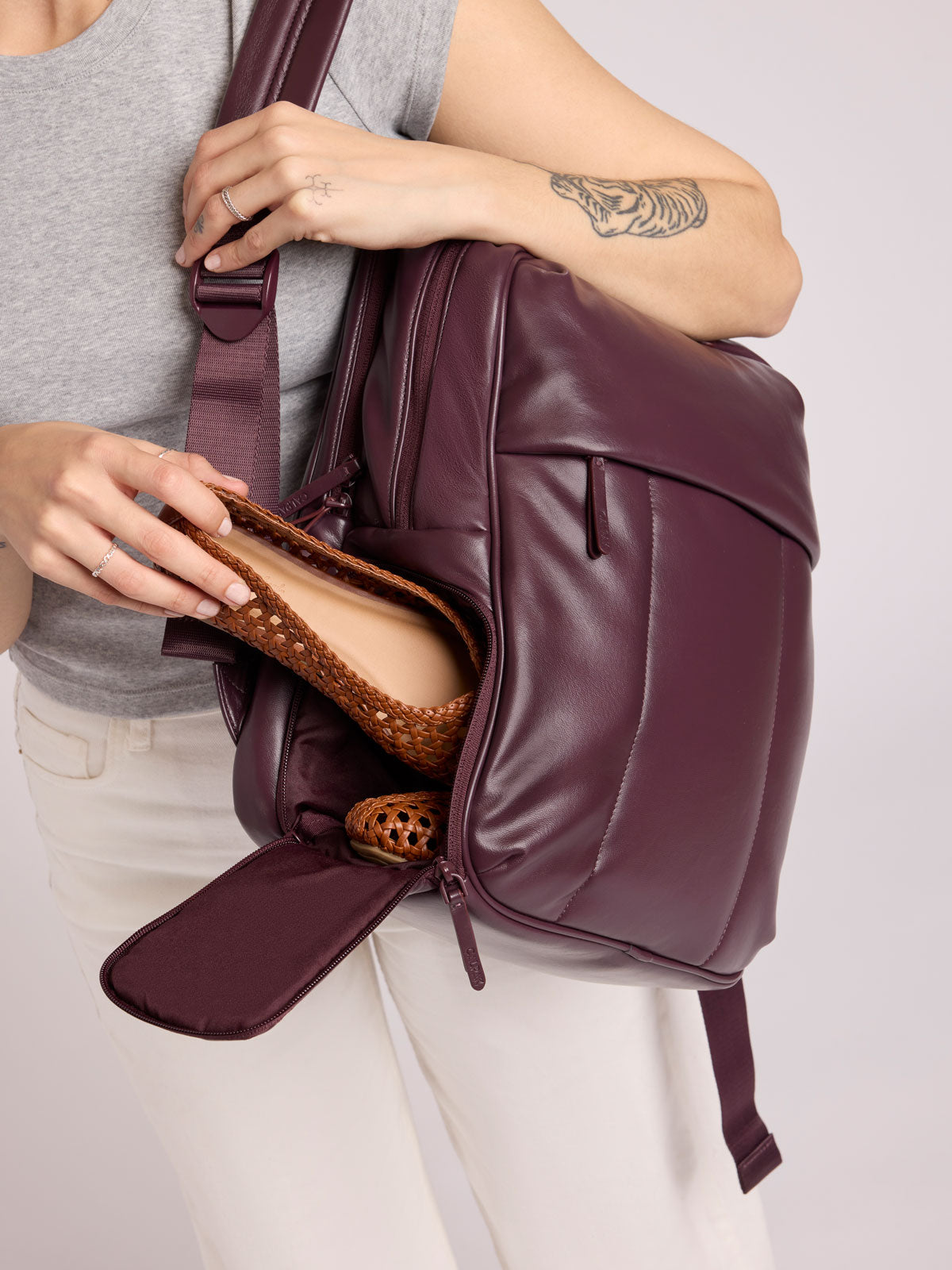 A person wearing a gray sleeveless top and white pants carries a CALPAK Luka 15in Luka Laptop Backpack in Dark Cherry Vegan Leather while putting in shoes in the bottom shoe compartment;  BPL2001-DARK-CHERRY-PU
