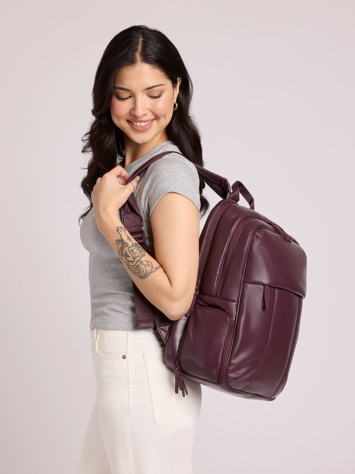 A woman with long dark hair, wearing a gray t-shirt and white pants, carries a CALPAK Luka 15in Laptop Backpack in Dark Cherry Vegan Leather over her shoulder against a light gray studio background;  BPL2001-DARK-CHERRY-PU