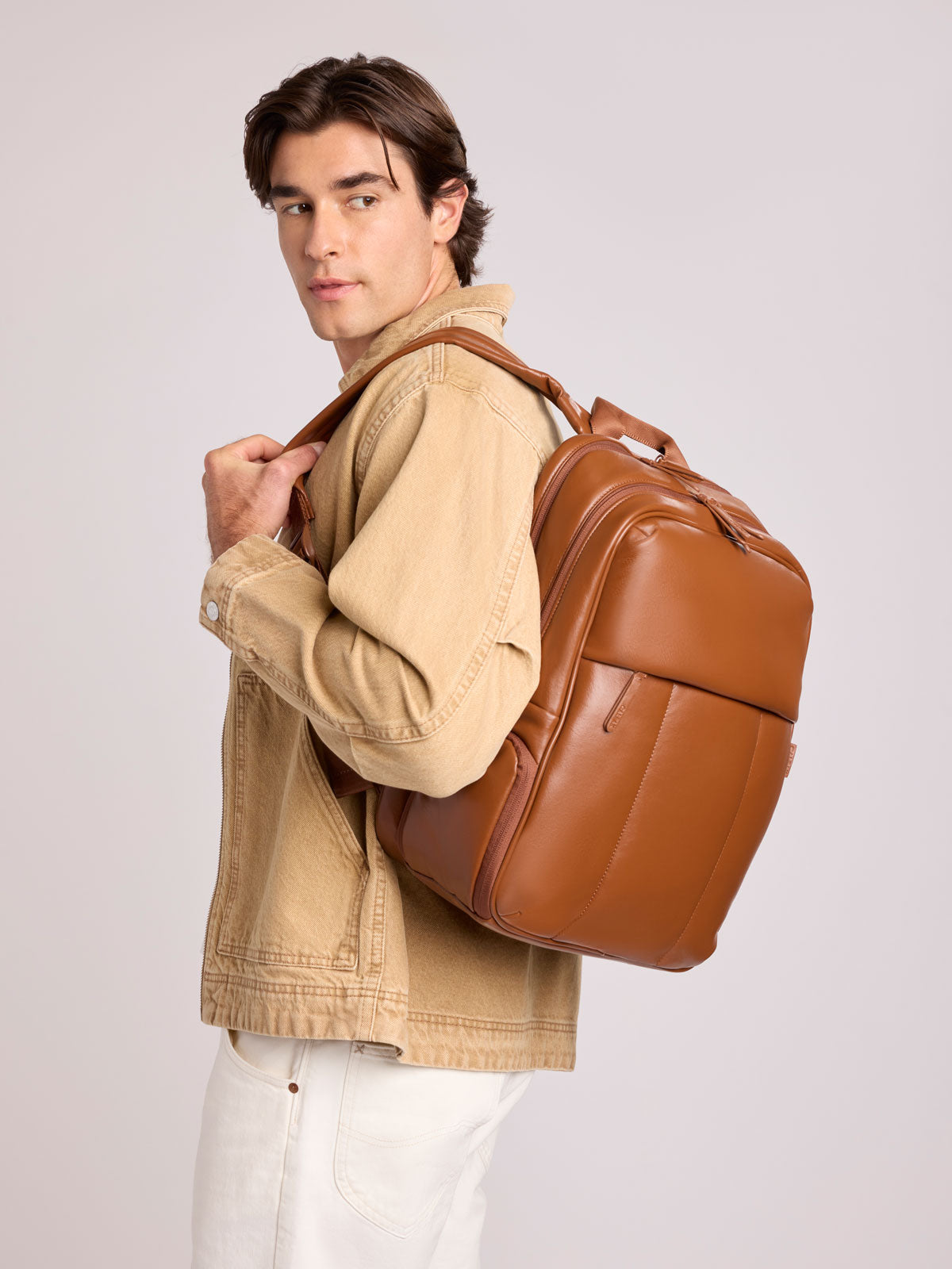 A man wearing a tan denim jacket and white pants carries a CALPAK Luka 15in Laptop Backpack in Honey Brown Vegan Leather over his shoulder, photographed against a light gray studio background; BPL2001-HONEY-BROWN-PU