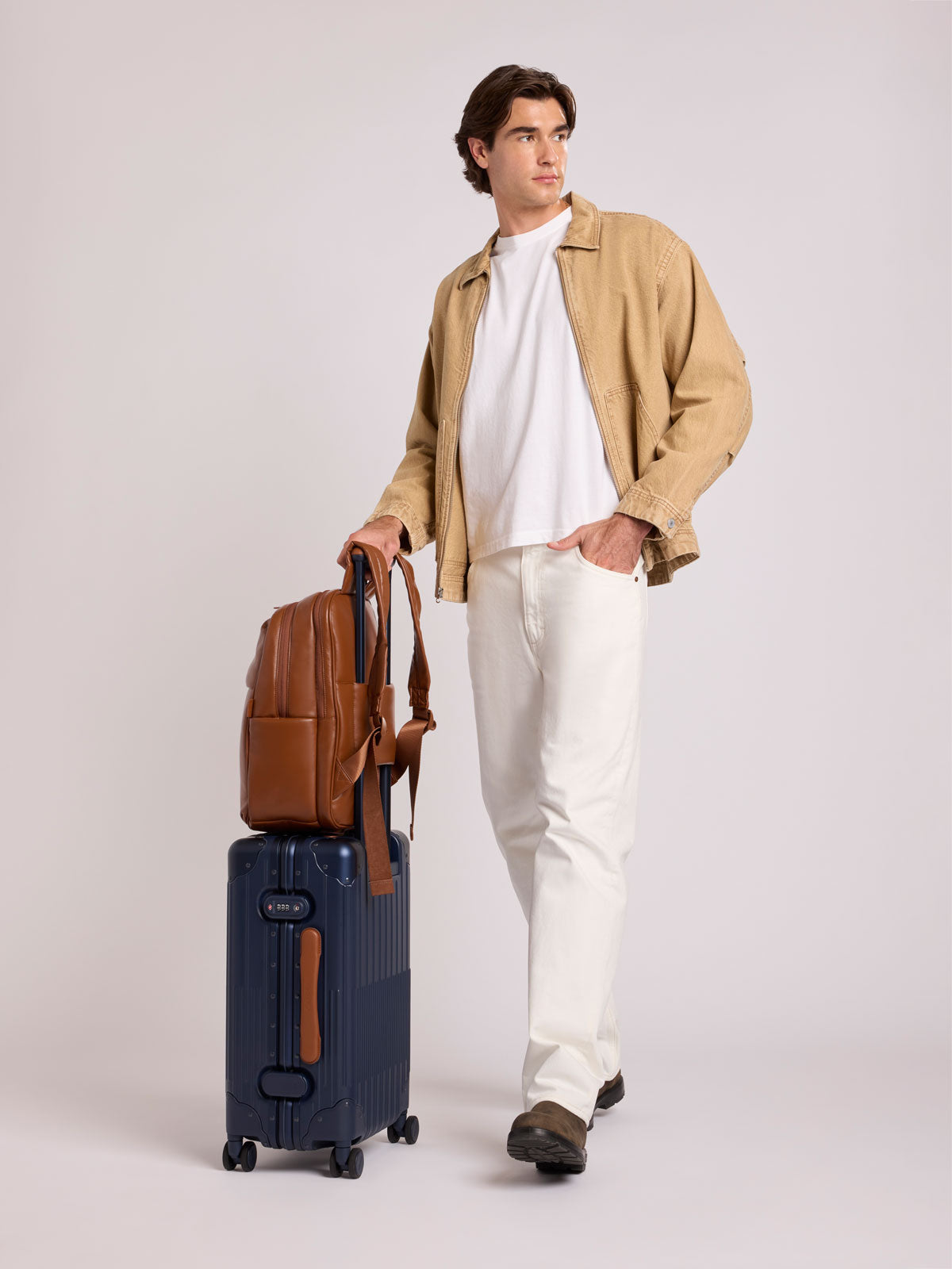A man stands in a studio wearing a beige overshirt, white T-shirt, and white trousers, holding a CALPAK Luka 15in Laptop Backpack in Honey Brown Vegan Leather on top of a navy Vault Carry On suitcase; BPL2001-HONEY-BROWN-PU
