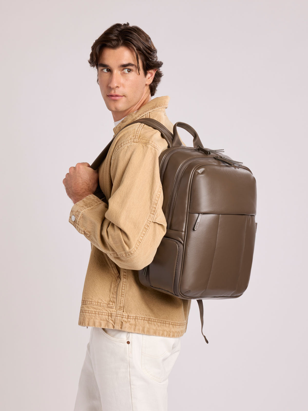A man wearing a tan jacket and white pants carries a CALPAK Luka 15in Laptop Backpack in Mushroom brown Vegan Leather on his shoulder, photographed against a light gray studio background; BPL2001-MUSHROOM-PU