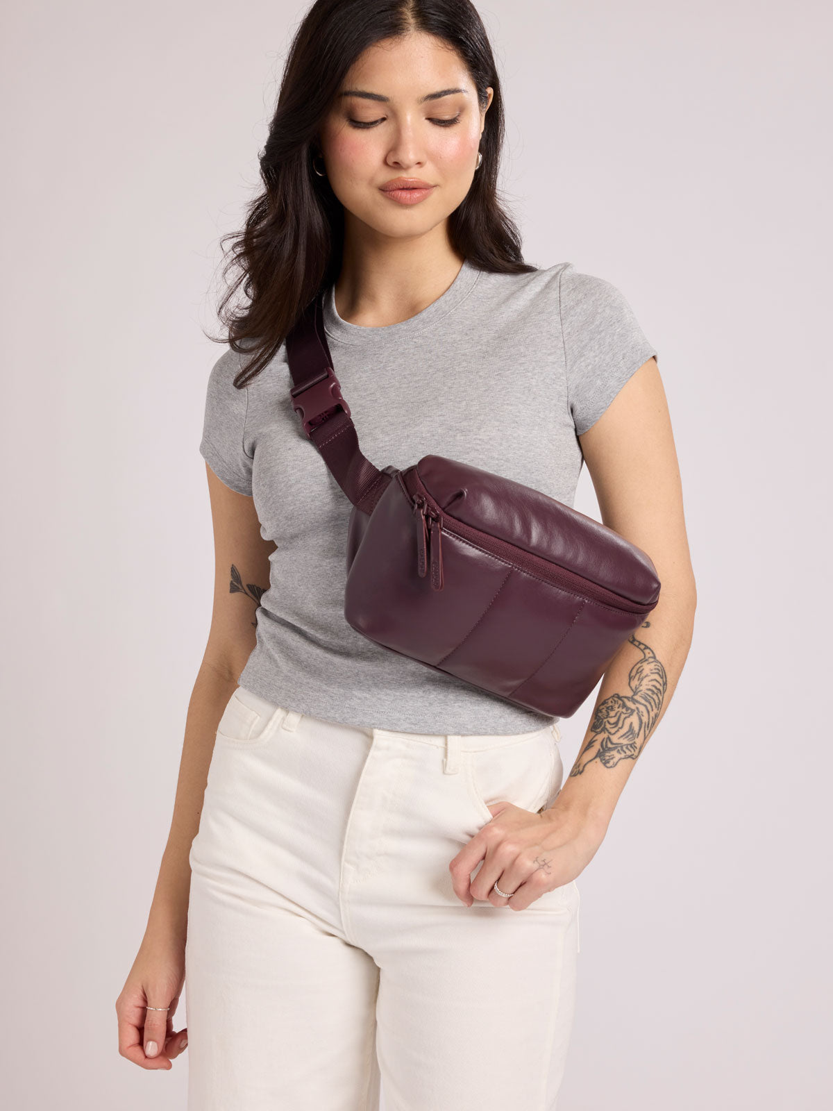 A woman wearing a heather gray t-shirt and white pants models a CALPAK Luka Belt Bag in Dark Cherry Vegan Leather  against a light gray studio background; BB1901-DARK-CHERRY-PU