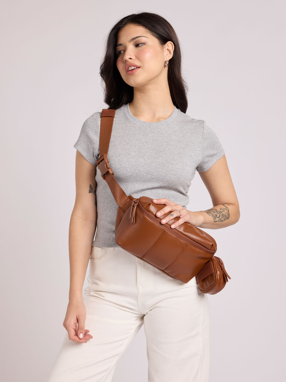 A woman wearing a light gray t-shirt and white pants models a CALPAK Luka Belt Bag in Honey Brown Vegan Leather, set against a solid light gray studio background; BB1901-HONEY-BROWN-PU
