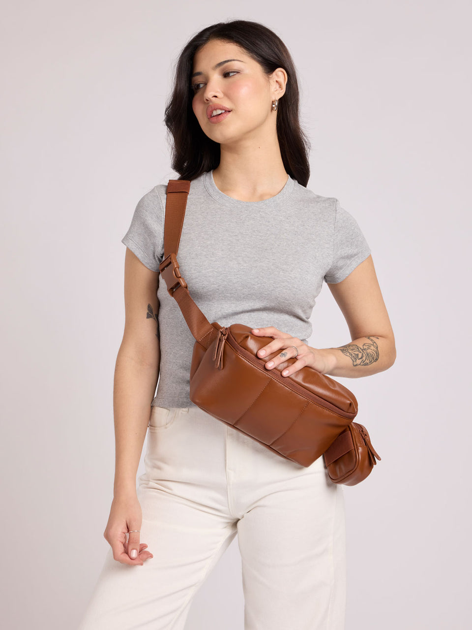 A woman wearing a light gray t-shirt and white pants models a CALPAK Luka Belt Bag in Honey Brown Vegan Leather, set against a solid light gray studio background; BB1901-HONEY-BROWN-PU
