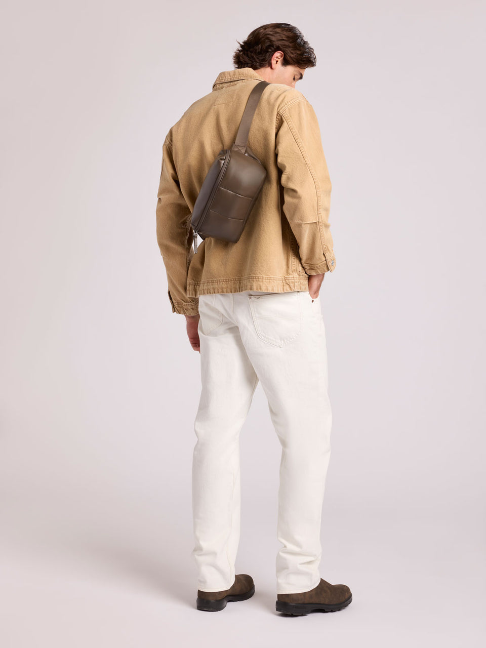 A man is shown from behind wearing a tan canvas utility jacket, white trousers, and brown shoes, with a CALPAK Luka Belt Bag in Mushroom brown Vegan Leather slung over his shoulder, set against a plain light gray studio background; BB1901-MUSHROOM-PU