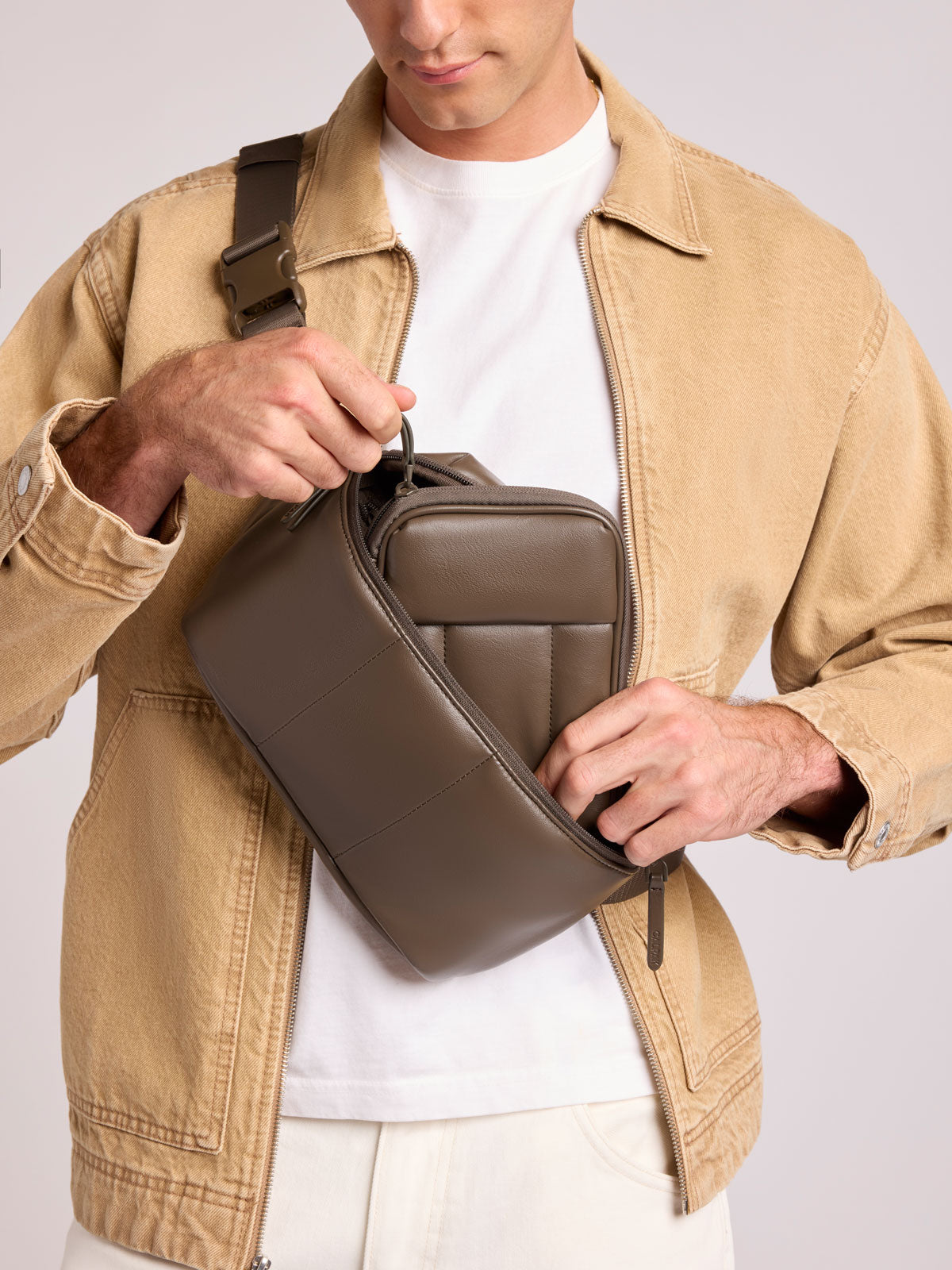 A man wearing a tan overshirt and white t-shirt models a CALPAK Luka Belt Bag in Mushroom Brown Vegan Leather with an adjustable strap, shown from the chest down against a light gray studio background; BB1901-MUSHROOM-PU