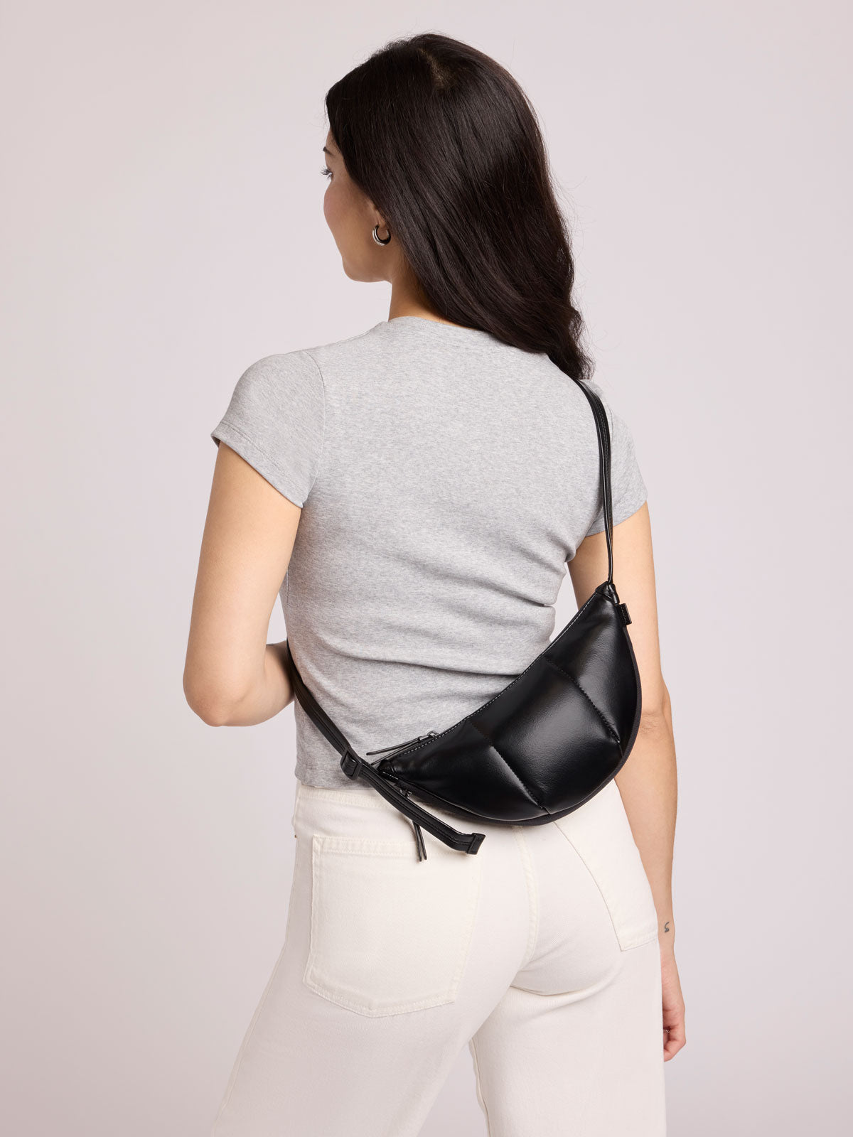 A woman shown from the back wearing a light gray t-shirt and white pants, carrying a CALPAK Luka Crescent Bag in Black Vegan Leather across her back; CBL2601-BLACK-PU