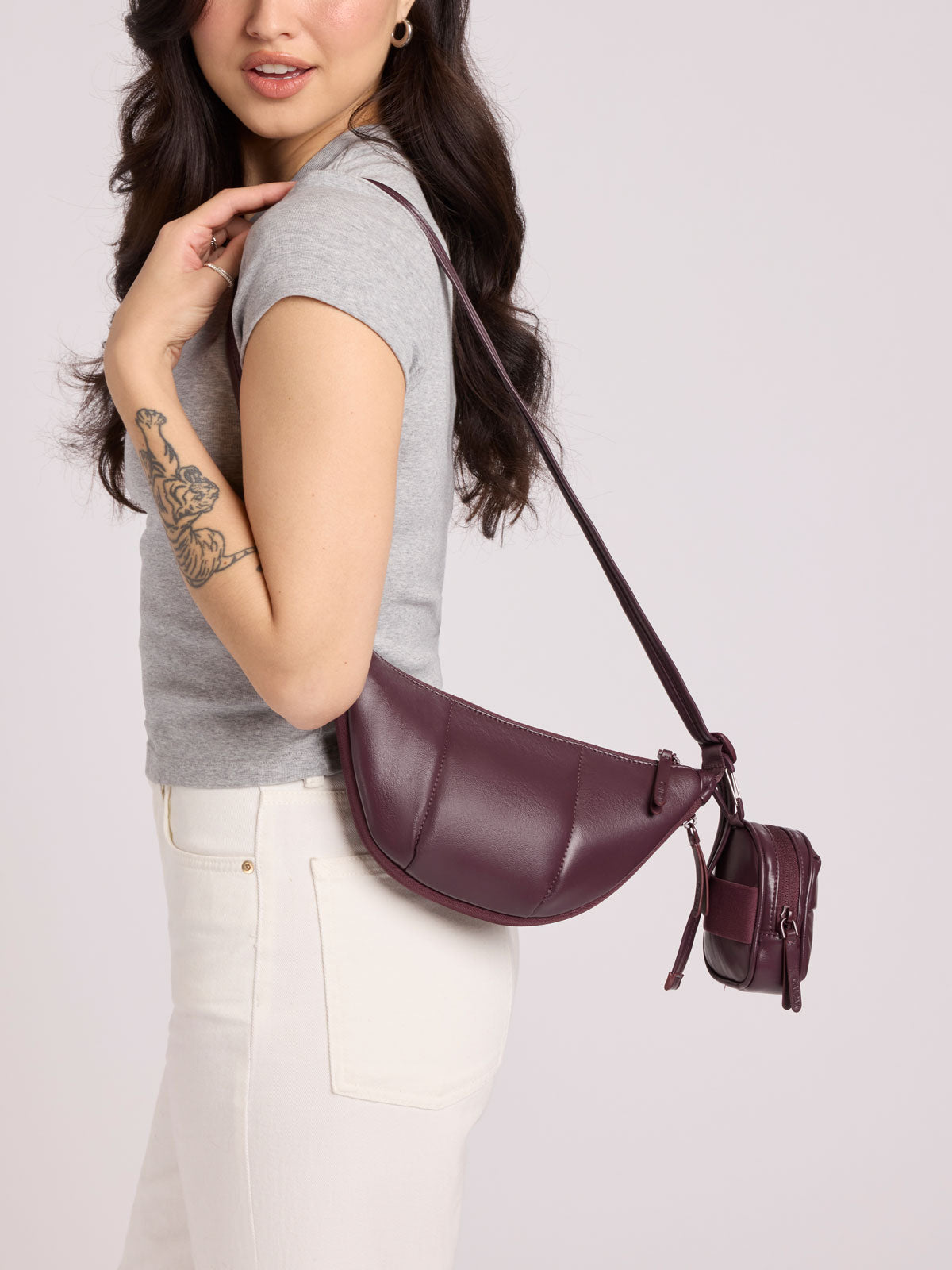 A woman wearing a gray t-shirt and white pants carries a Luka Crescent Bag with the Luka Backpack Zip Bag Charm attached in Dark Cherry Vegan Leather against a light gray studio background; CBL2601-DARK-CHERRY-PU