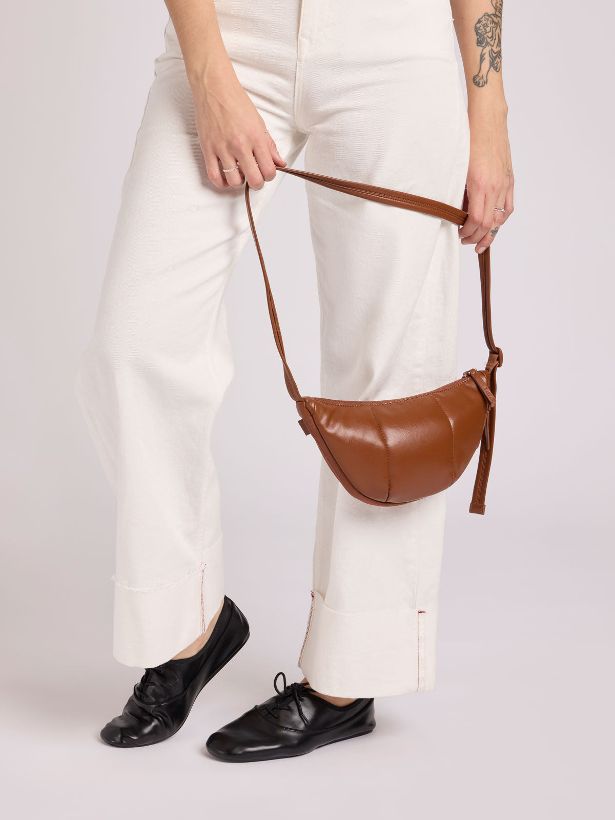 A person wearing white wide-leg trousers and black loafers holds a CALPAK Luka Crescent Bag in Honey Brown Vegan Leather with a long strap against a light gray studio background; CBL2601-HONEY-BROWN-PU